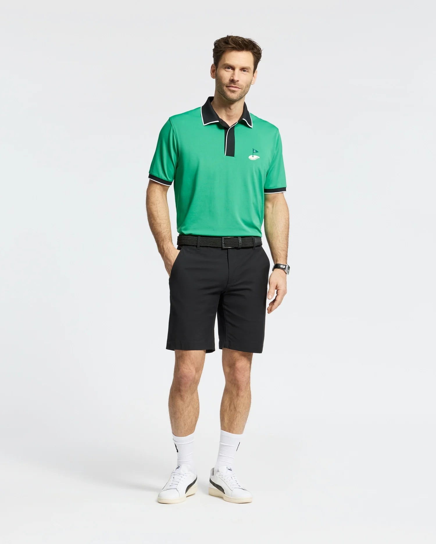 A man in a Psycho Bunny MENS CLAYTON PIQUE POLO - MT0100004, made of breathable Pima cotton with green and black colorblocked details and a small emblem, stands against a white background. He's wearing black shorts, white sneakers, and sports a watch on his left wrist with one hand in his pocket.