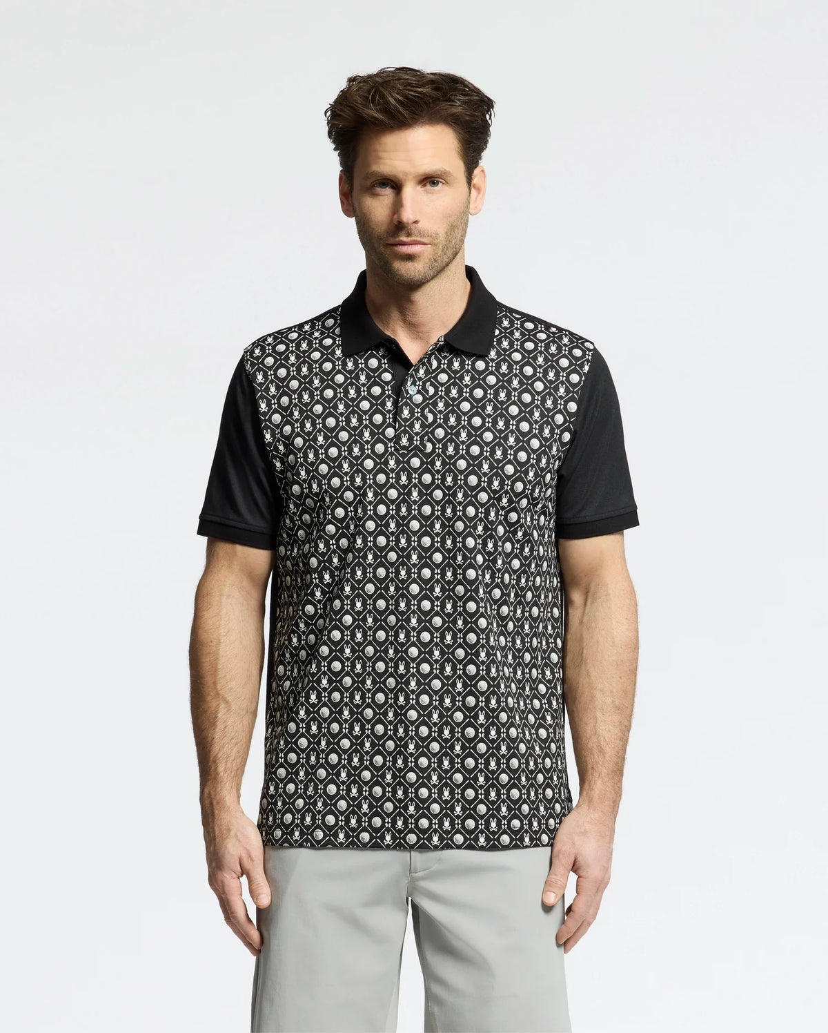 A person in a stylish Psycho Bunny Men's Beckett Pique Polo (MT0100022) featuring a black and white pattern with black sleeves stands against a white background, paired with light gray pants that enhance their short, styled hair.