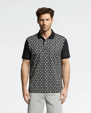 A person in a stylish Psycho Bunny Men's Beckett Pique Polo (MT0100022) featuring a black and white pattern with black sleeves stands against a white background, paired with light gray pants that enhance their short, styled hair.
