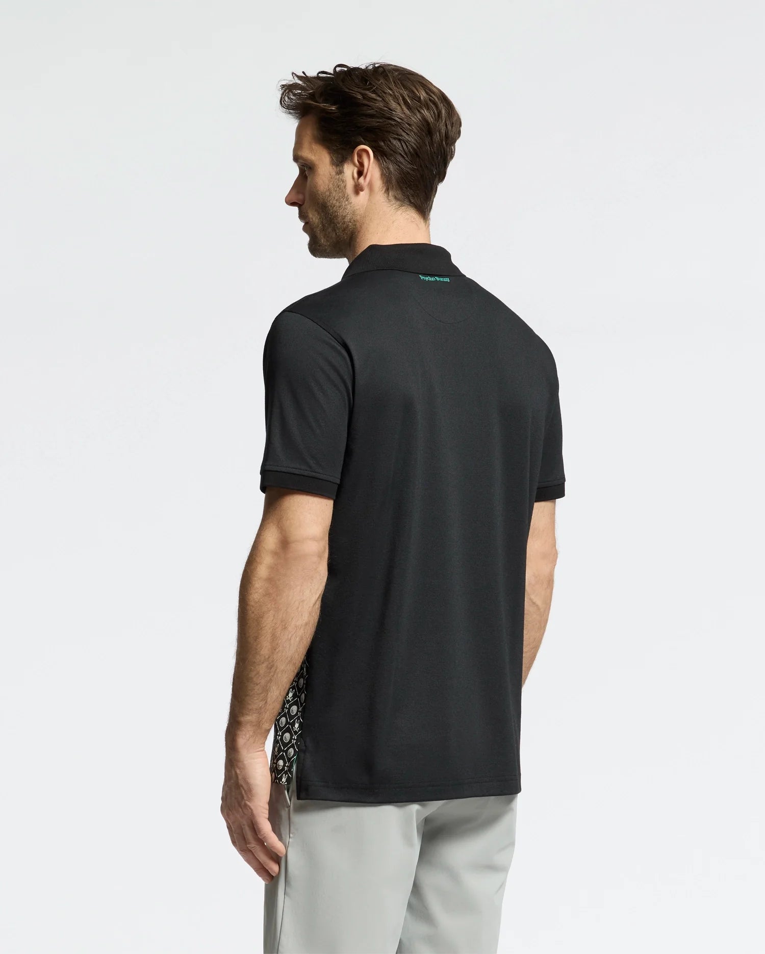 A man stands in profile, gazing left, dressed in a black MENS BECKETT PIQUE POLO - MT0100022 by Psycho Bunny, made with breathable fabric and featuring a textured side pattern. He's also wearing light gray pants against a neutral background.