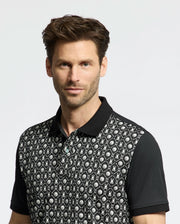A man with short brown hair wears the Psycho Bunny Men's Beckett Pique Polo (MT0100022), featuring a geometric and leaf pattern in black. The background is plain and light-colored.