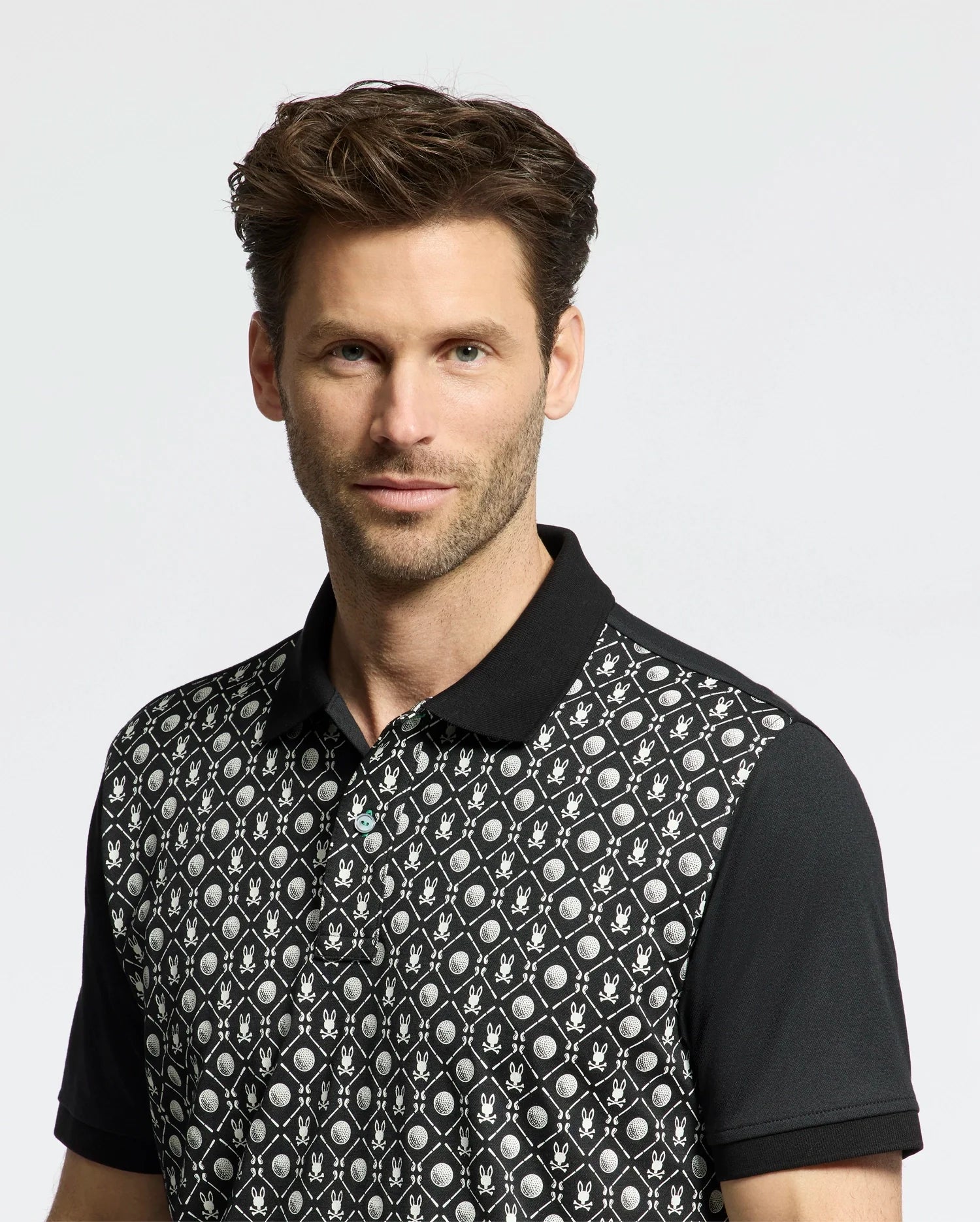 A man with short brown hair wears the Psycho Bunny Men's Beckett Pique Polo (MT0100022), featuring a geometric and leaf pattern in black. The background is plain and light-colored.