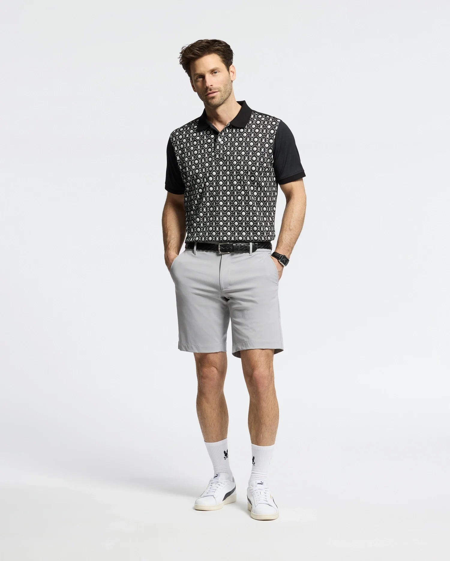 A person stands against a plain background wearing a Psycho Bunny MENS BECKETT PIQUE POLO (MT0100022) in black, light gray shorts, white socks, and white sneakers. With hands in pockets and neatly styled hair, the breathable polo adds an effortless touch to their ensemble.