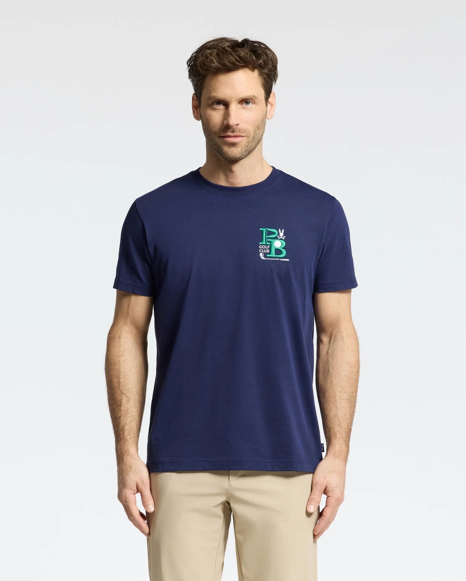 A man with short brown hair wears the navy blue Psycho Bunny MENS DENIS GRAPHIC TEE - MT0200015, made from soft Pima cotton with a small embroidered logo on the upper left. He pairs it with beige pants for a golf-inspired look, standing against a plain white background.