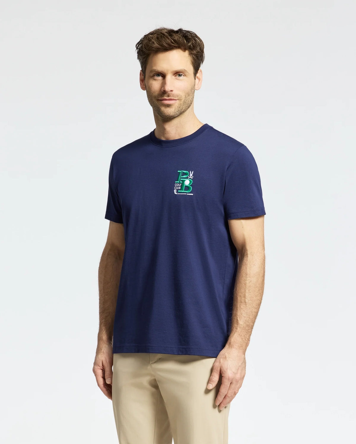 A man with short brown hair wears a Psycho Bunny MENS DENIS GRAPHIC TEE in navy blue, featuring a small embroidered logo on the chest, along with beige pants. The golf-inspired look stands out against the plain light background as he faces forward with a relaxed expression.
