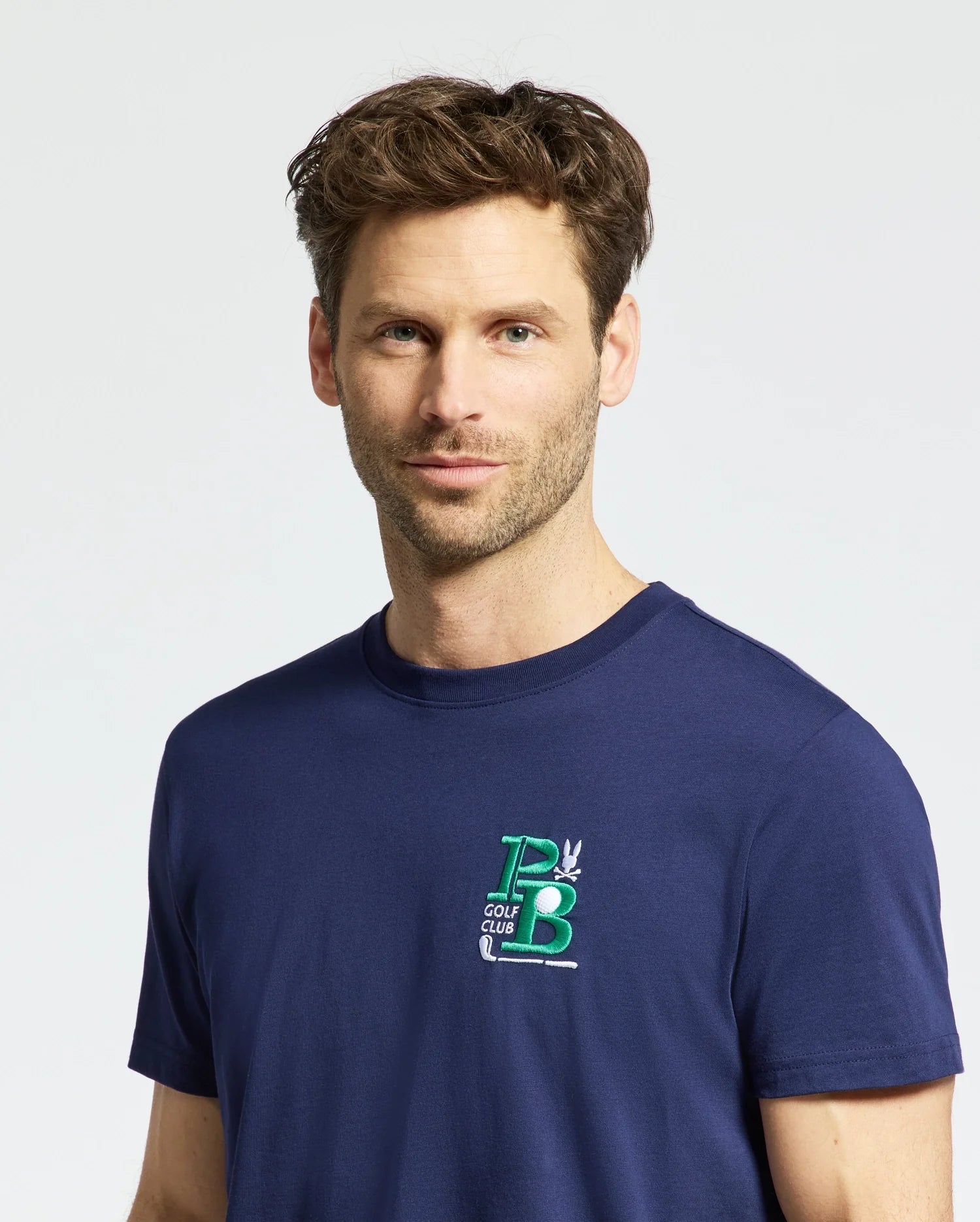 A man with brown hair wears a Psycho Bunny MENS DENIS GRAPHIC TEE - MT0200015, a navy blue Pima cotton shirt with an embroidered green and white "PB Golf Club" logo. He stands against a light background, gazing slightly to the side in golf-inspired style.