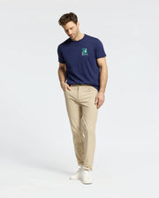 A man is wearing a Psycho Bunny navy MENS DENIS GRAPHIC TEE featuring an embroidered "E" on the chest, paired with beige pants and white sneakers. He stands against a plain white background, looking down with hands in his pockets.