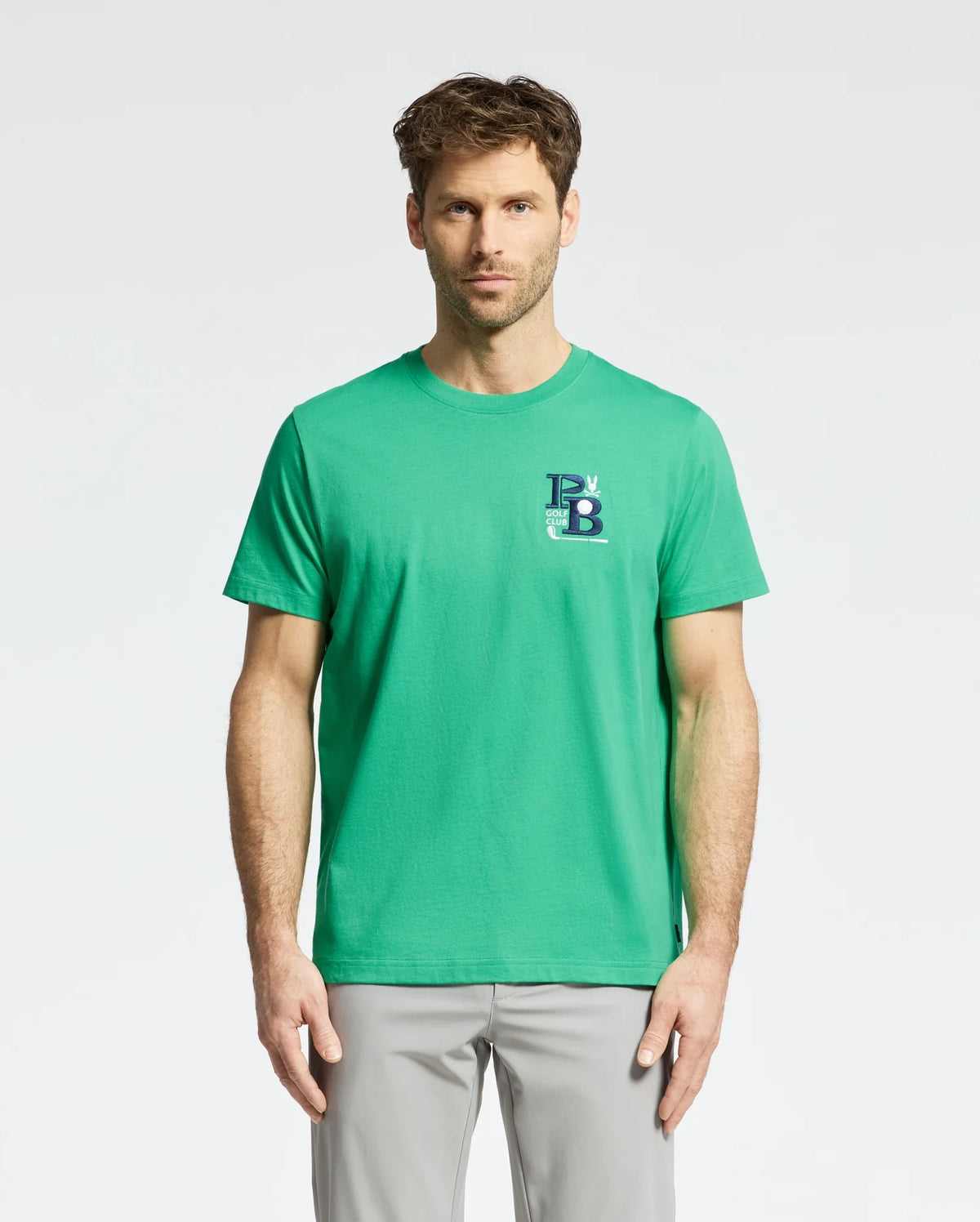 A man wearing a Psycho Bunny Men's Denis Graphic Tee - MT0200015 in green, featuring an embroidered logo on the left side, and light gray pants stands against a plain background.