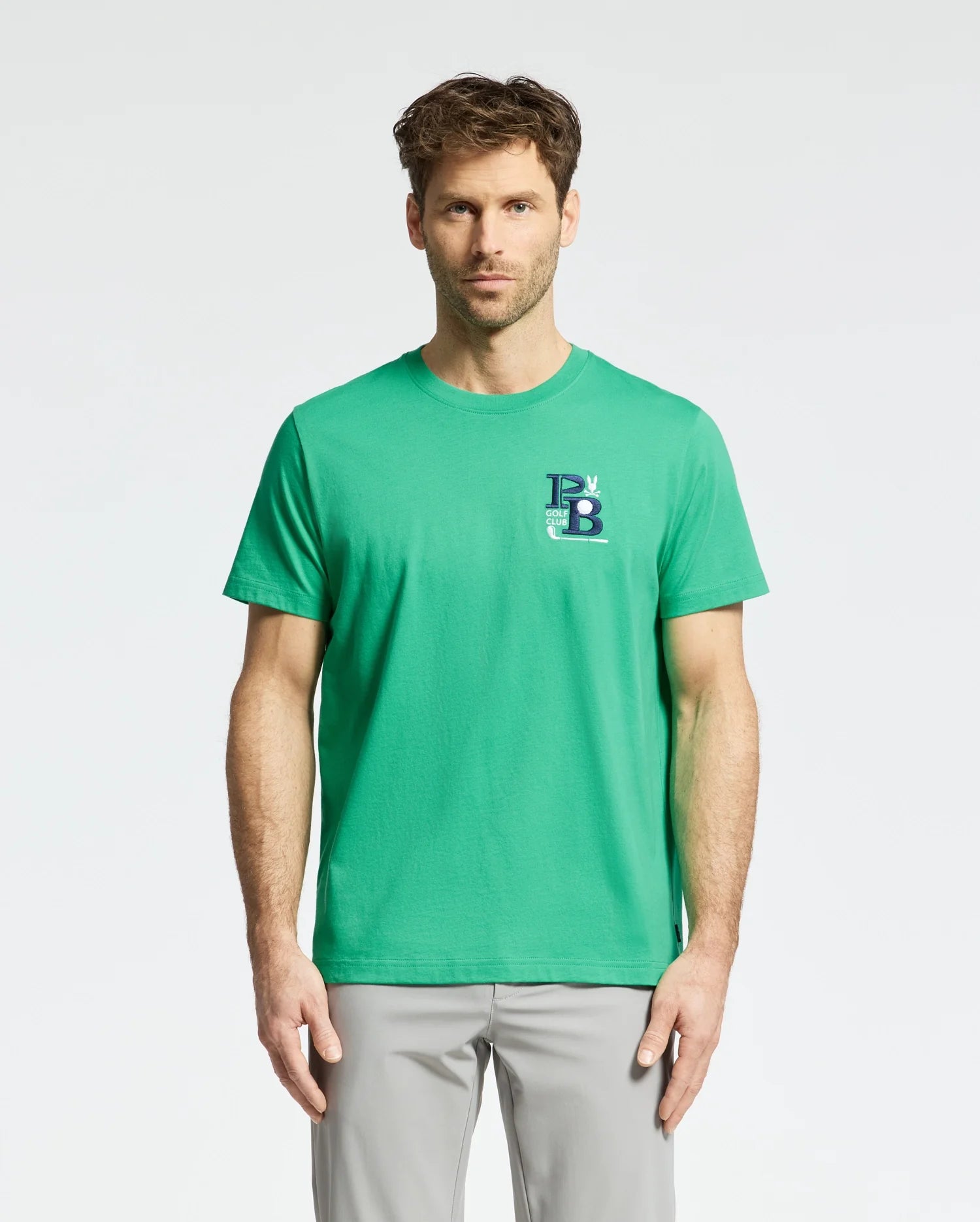 A man wearing a Psycho Bunny Men's Denis Graphic Tee - MT0200015 in green, featuring an embroidered logo on the left side, and light gray pants stands against a plain background.