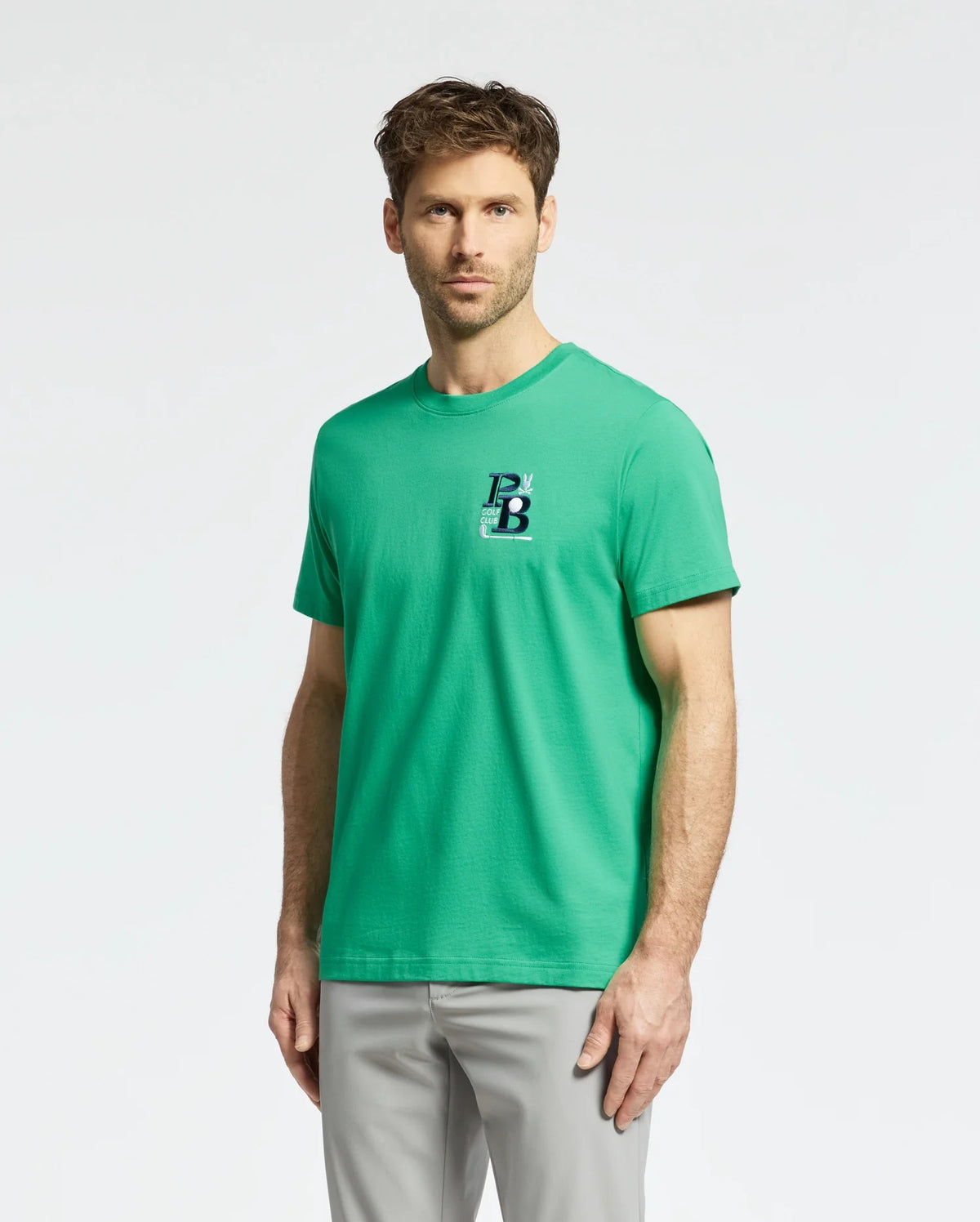 A man with short, wavy brown hair and a trimmed beard wears the Psycho Bunny MENS DENIS GRAPHIC TEE (MT0200015), showcasing its "PB" logo on the chest. Made from Pima cotton, this green tee complements his light gray pants, giving off a casual, golf-inspired vibe against a plain background.