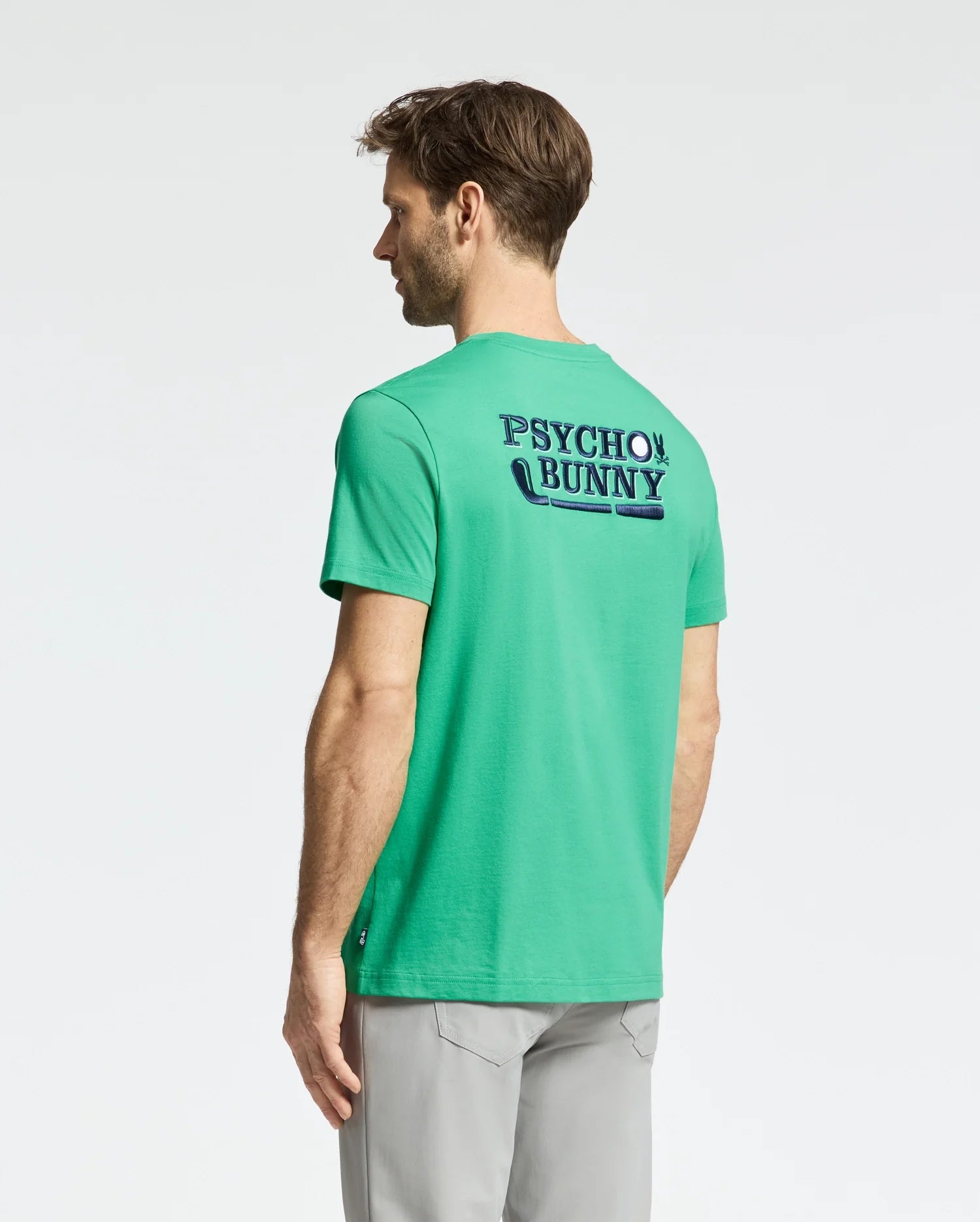 A man with short hair faces away, wearing a bright green MENS DENIS GRAPHIC TEE - MT0200015 by Psycho Bunny, featuring "Psycho Bunny" in bold print and an embroidered logo. Light gray pants complete his look against the plain white backdrop.