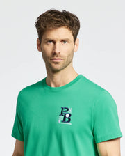 A person with short, wavy hair stands against a plain white background wearing the MENS DENIS GRAPHIC TEE - MT0200015 by Psycho Bunny. The green Pima cotton T-shirt features an embroidered logo on the chest. Their expression is neutral.