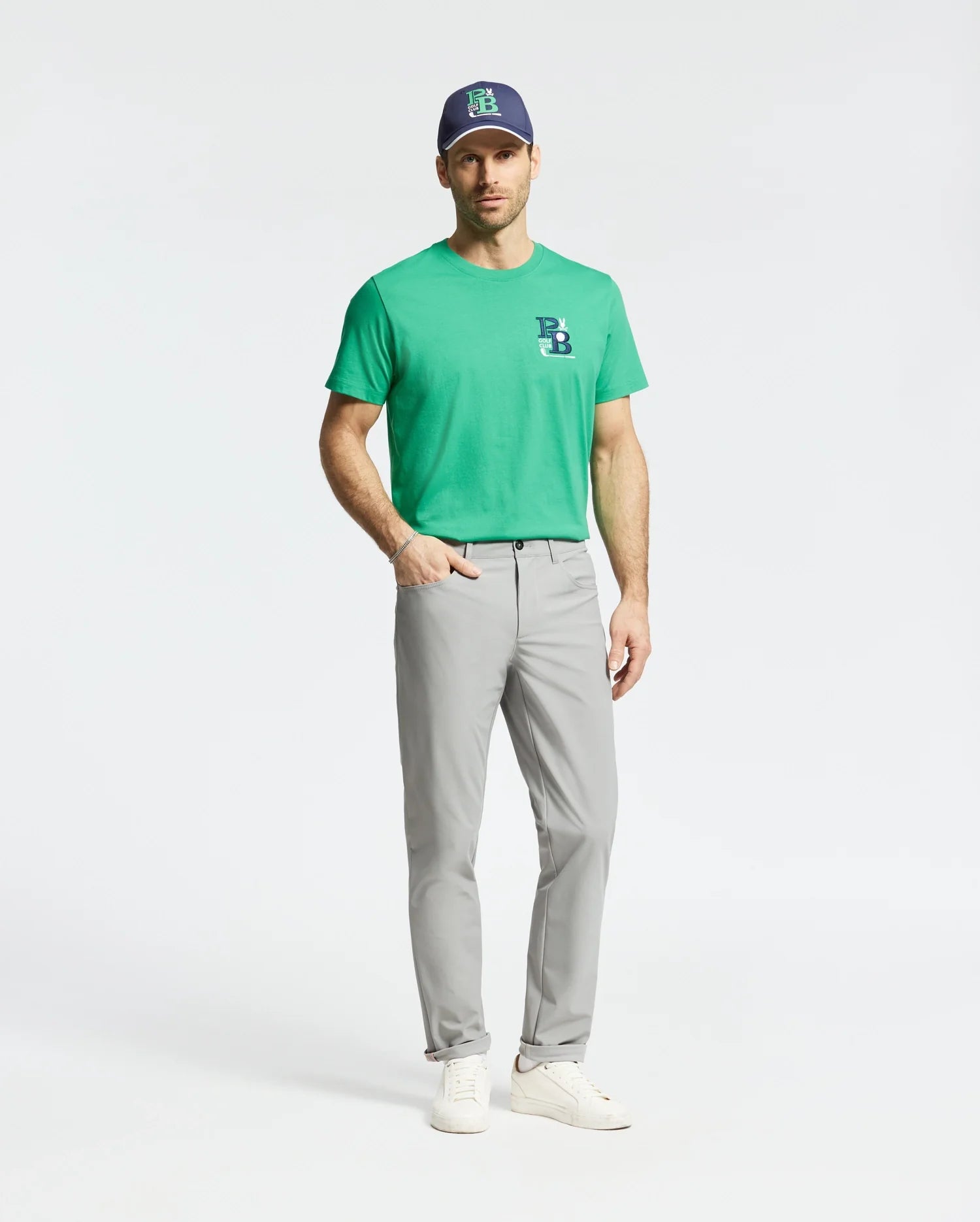 A man stands against a plain background wearing Psycho Bunny's MENS DENIS GRAPHIC TEE (MT0200015) in golf-inspired green, crafted from soft Pima cotton with an embroidered logo, light gray pants, a navy cap featuring a green logo, and white sneakers.