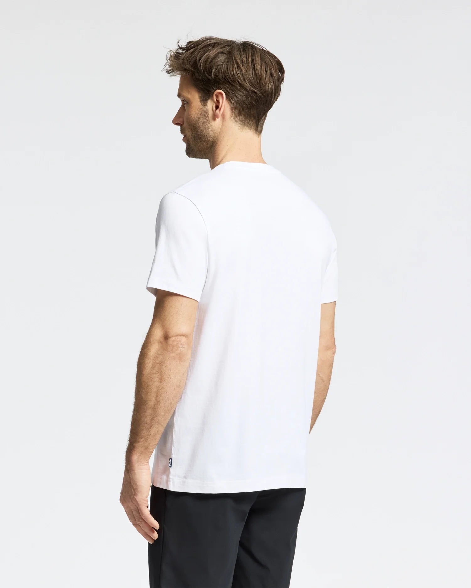 A man with short brown hair faces away, wearing a Psycho Bunny MENS DALE GRAPHIC TEE - MT0200016 and black pants. The light neutral background highlights the simplicity of his casual style.