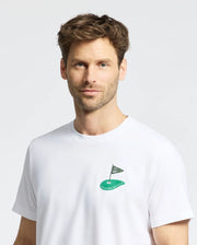 A man with short brown hair and a beard is wearing the Mens Dale Graphic Tee (MT0200016) from Psycho Bunny. It's a Pima cotton white t-shirt featuring an embroidered golf hole and flag logo on the chest, with "Psycho Bunny" text. He stands against a light gray background.