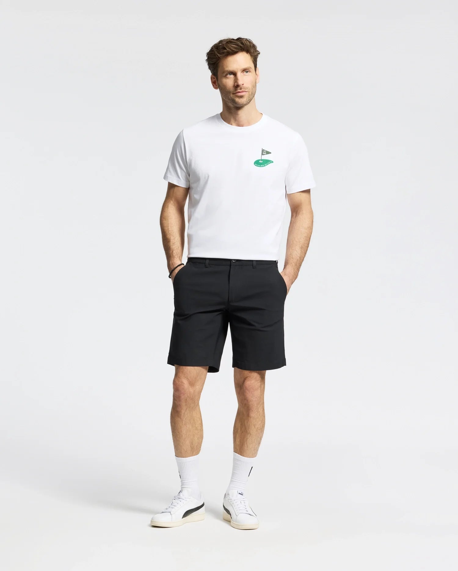 A man in the Psycho Bunny MENS DALE GRAPHIC TEE (MT0200016), a white Pima cotton T-shirt with a green embroidered logo, pairs it with black shorts. He wears white sneakers and socks, posing against a plain background with understated yet refined casual elegance.