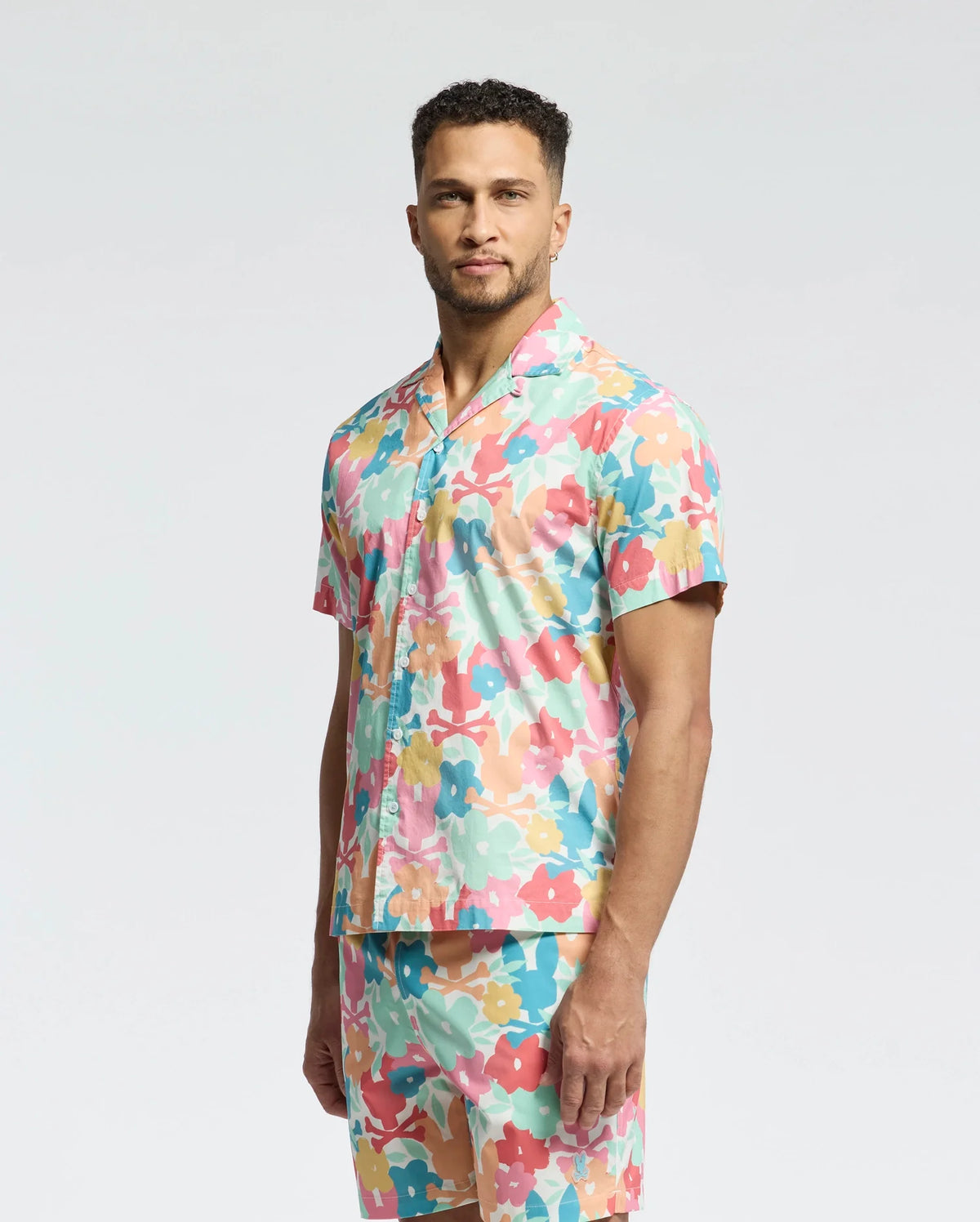 A man stands confidently against a plain background wearing the Psycho Bunny MENS MEYLON STRETCH POPLIN SHIRT (MT0300034) and matching pants. The ensemble features a vibrant floral pattern in pink, green, orange, and blue with a camp collar that complements his short curly hair and beard.