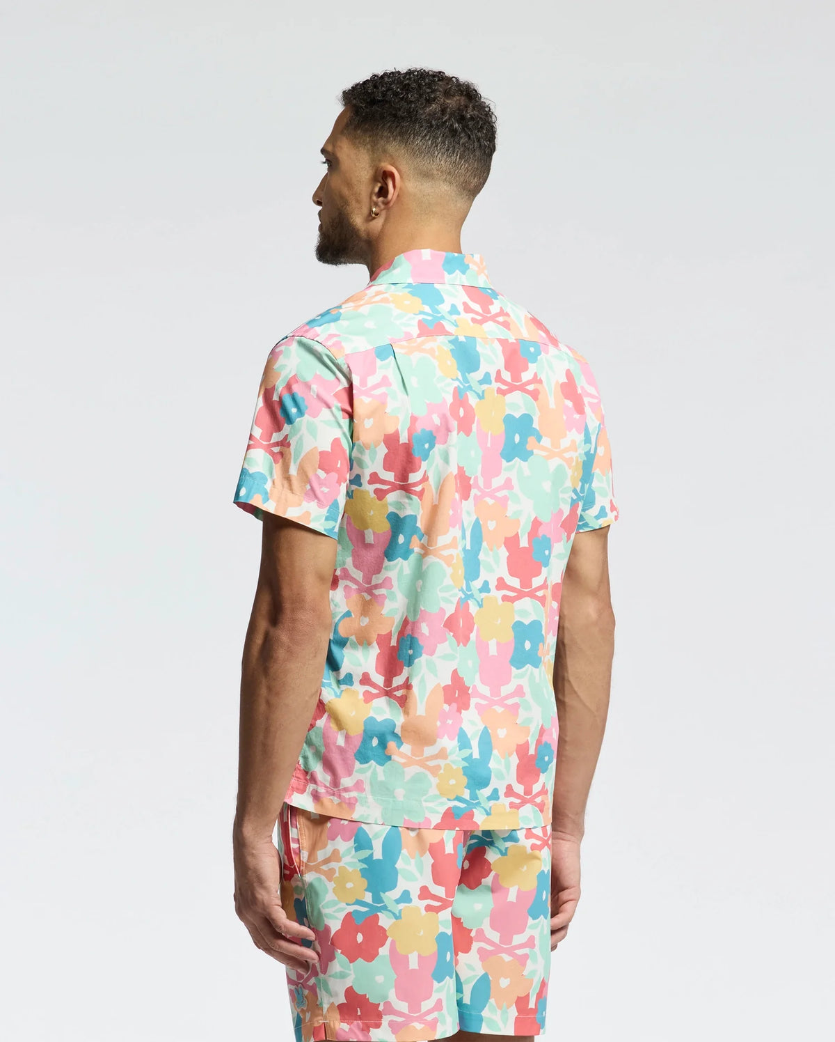 A man with short curly hair stands with his back to the camera, wearing Psycho Bunny's MENS MEYLON STRETCH POPLIN SHIRT (MT0300034) featuring a colorful floral camp collar and matching shorts against a plain white background.