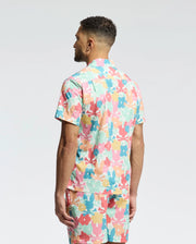 A man with short curly hair stands with his back to the camera, wearing Psycho Bunny's MENS MEYLON STRETCH POPLIN SHIRT (MT0300034) featuring a colorful floral camp collar and matching shorts against a plain white background.