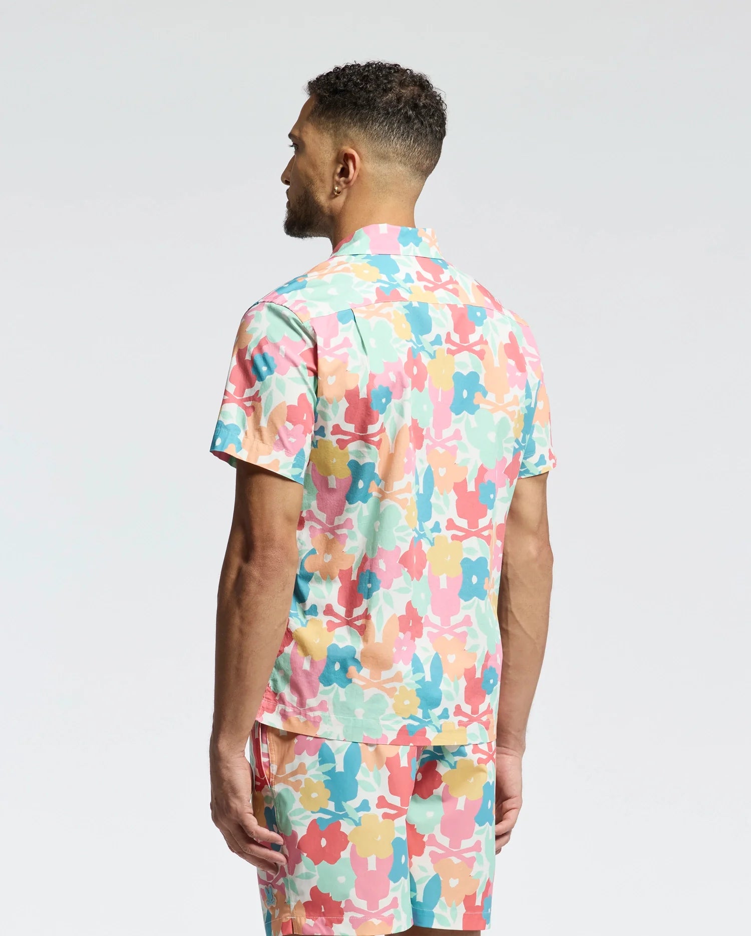 A man with short curly hair stands with his back to the camera, wearing Psycho Bunny's MENS MEYLON STRETCH POPLIN SHIRT (MT0300034) featuring a colorful floral camp collar and matching shorts against a plain white background.