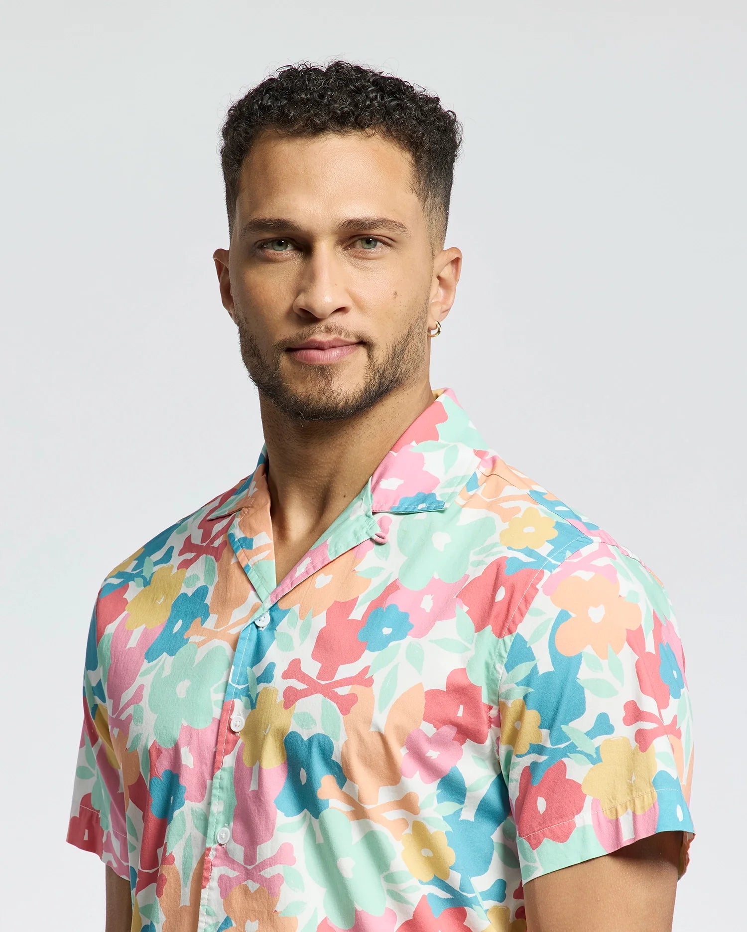 A person with short curly hair wears Psycho Bunny's MENS MEYLON STRETCH POPLIN SHIRT - MT0300034 against a plain background, gazing at the camera with a slight smile.