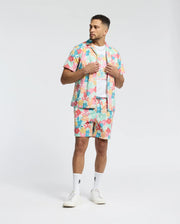 A man wears a Psycho Bunny MENS MEYLON STRETCH POPLIN SHIRT (MT0300034) with matching shorts, adorned in abstract patterns and a camp collar. With a white t-shirt beneath, white socks, and sneakers, he stands against a plain background, hands on his open shirt, looking to the side.