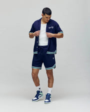A man wears the Psycho Bunny MENS BASIL TAPED CAMP COLLAR SHIRT (MT0500004) in navy, paired with matching shorts, a white tank top, white socks, and blue-green sneakers. He looks down while adjusting his shirt against a plain background.
