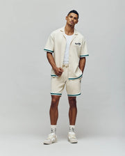 A man stands against a plain background wearing the Psycho Bunny MENS BASIL TAPED CAMP COLLAR SHIRT (MT0500004), matching shorts with green trim, a white tank, white socks, and cream sneakers. One hand is in his pocket.