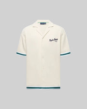 The Psycho Bunny MENS BASIL TAPED CAMP COLLAR SHIRT (MT0500004) is cream-colored, made of a cotton-modal blend with short sleeves, green/blue stripe accents, a Johnny collar, and embroidered "Psycho Bunny" text on the left chest.