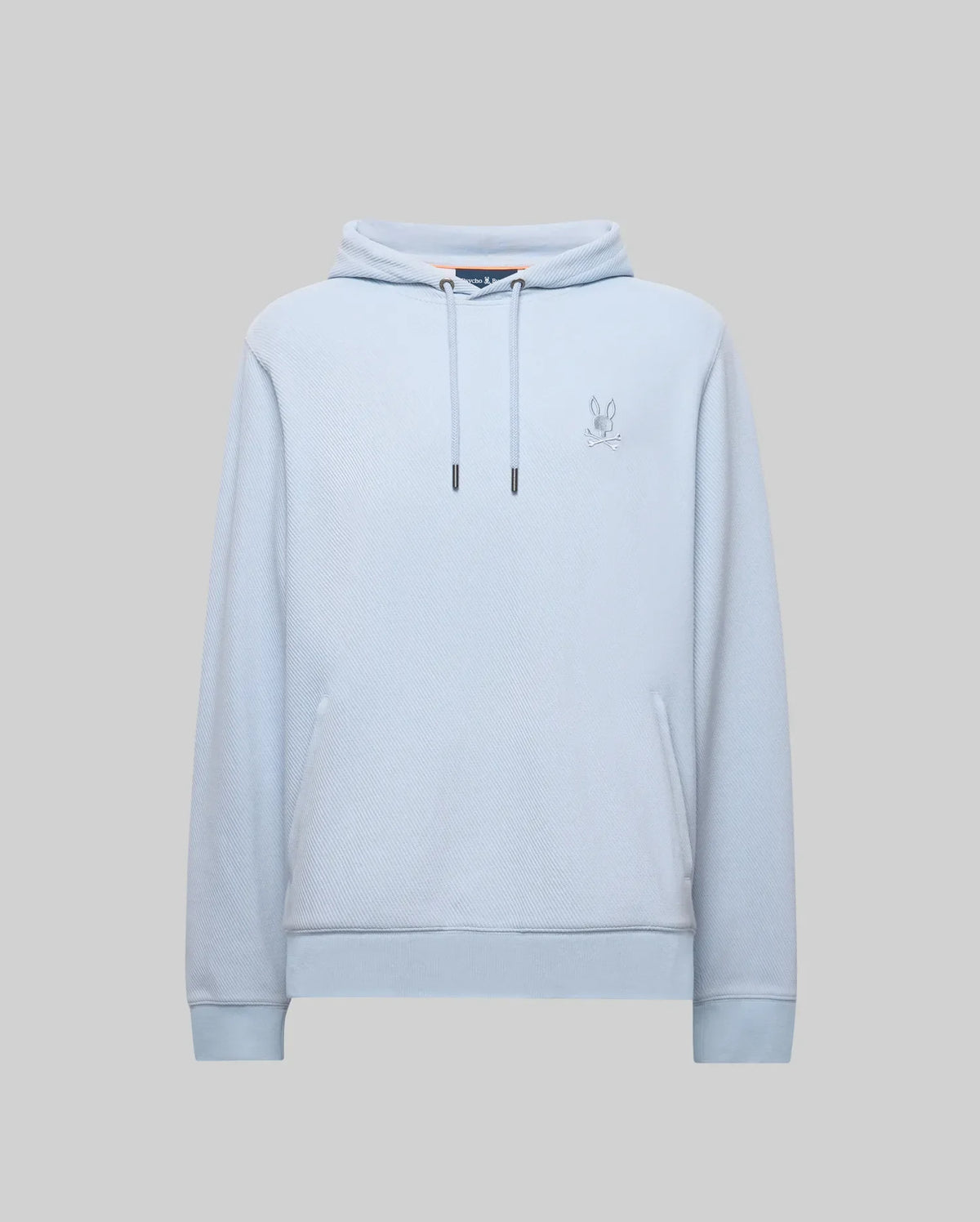 The Psycho Bunny MENS KAI HOODIE - MT0500019 features a light blue twill texture, front pocket, drawstrings, and an embroidered Bunny logo on the left chest, shown against a gray background.