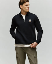 A man in a black Psycho Bunny MENS DANIEL QUARTER ZIP SWEATSHIRT (MT0500023) with a bunny logo, white shirt underneath, and olive green cargo pants stands against a plain light gray background.