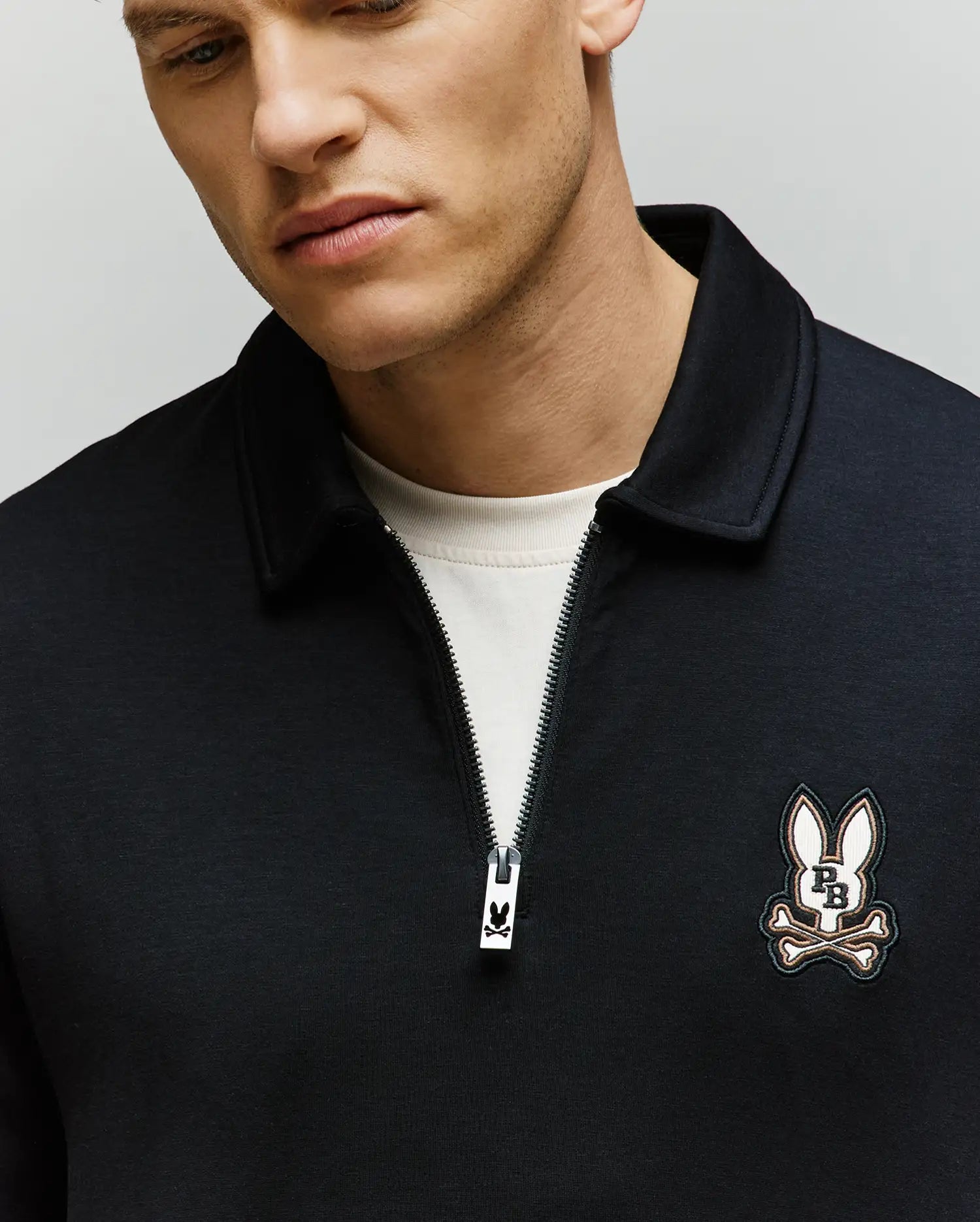 A man wears the Psycho Bunny MENS DANIEL QUARTER ZIP SWEATSHIRT (MT0500023) over a white T-shirt, showing the bunny and crossbones patch on the chest. His face is partially visible as he looks down slightly.