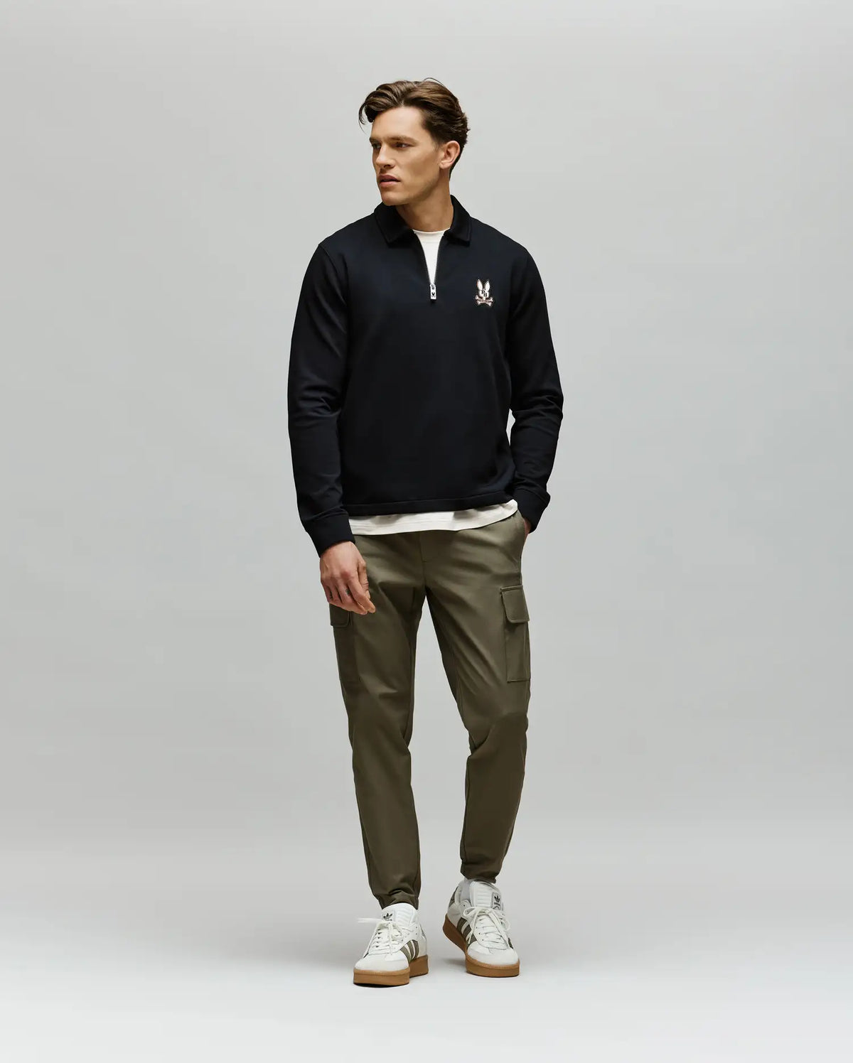 A man stands against a plain gray background, wearing a Psycho Bunny MENS DANIEL QUARTER ZIP SWEATSHIRT - MT0500023 in black, olive green cargo joggers, and white sneakers. He looks to his left with one hand in his pocket.