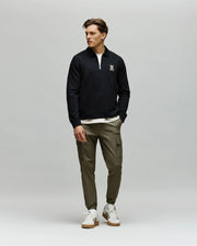 A man stands against a plain gray background, wearing a Psycho Bunny MENS DANIEL QUARTER ZIP SWEATSHIRT - MT0500023 in black, olive green cargo joggers, and white sneakers. He looks to his left with one hand in his pocket.