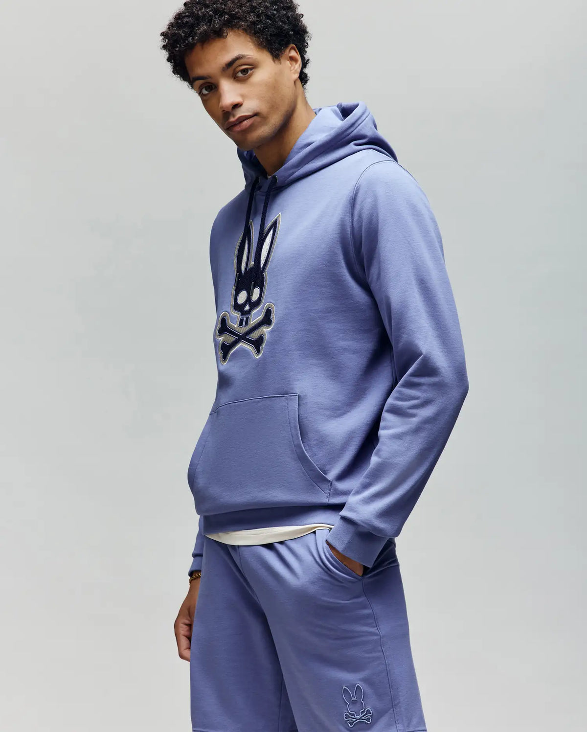 A man wears the Psycho Bunny MENS RYDER BUNNY HOODIE (MT0500029) in light purple with matching shorts, featuring a black and white embroidered Bunny patch. He stands against a plain background, looking slightly to the side with one hand in his pocket.