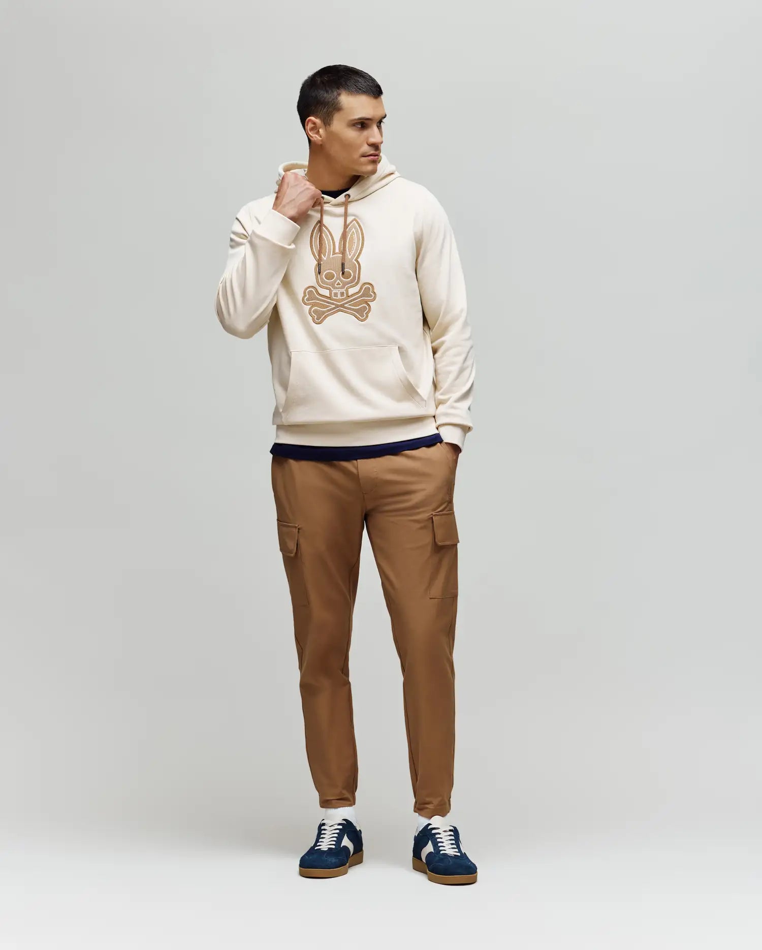 A man stands against a plain background, wearing the Psycho Bunny MENS RYDER BUNNY HOODIE (MT0500029) in cream, paired with tan cargo pants and navy sneakers with white soles for a relaxed loungewear look.