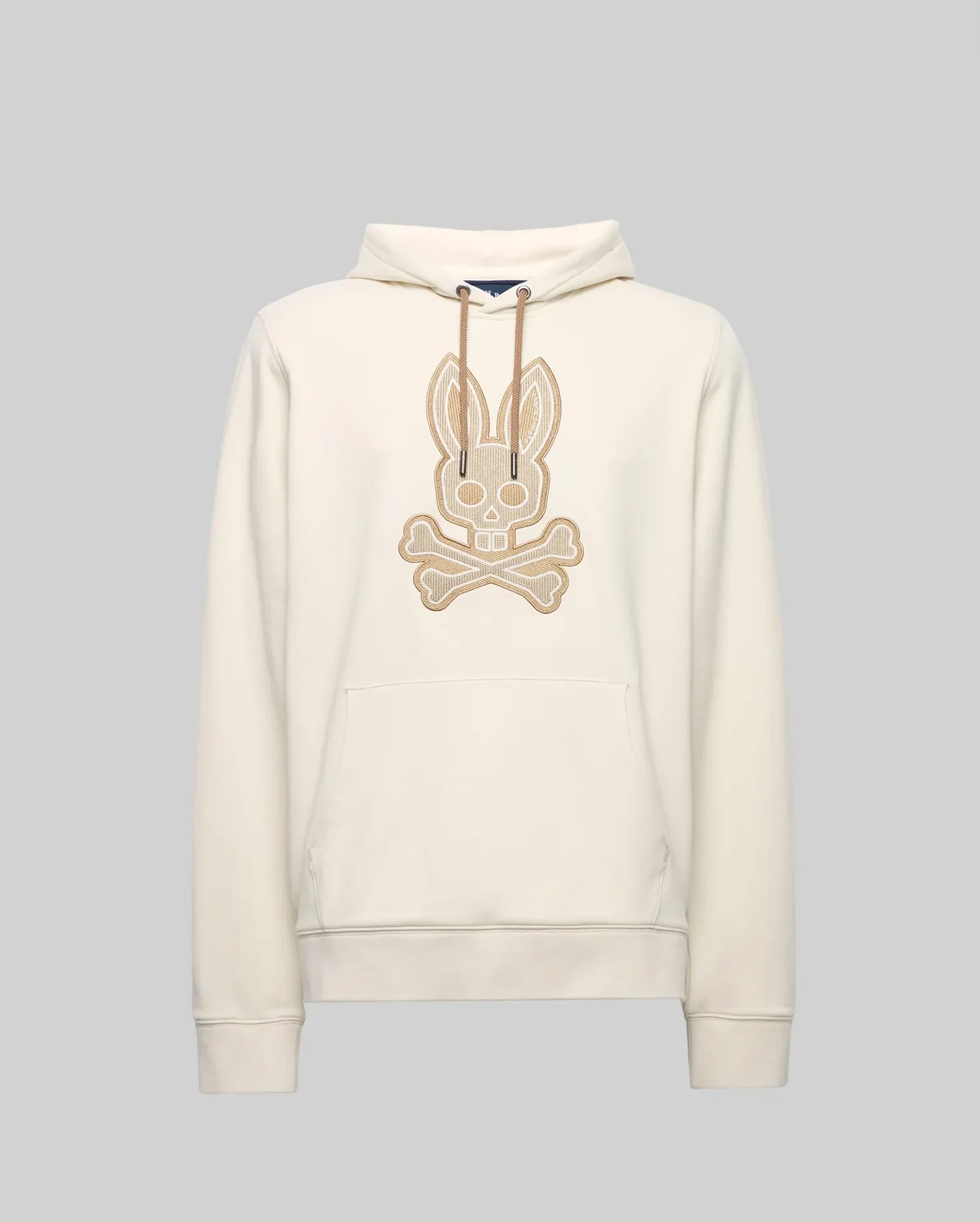 The Psycho Bunny MENS RYDER BUNNY HOODIE (MT0500029) is a cream hoodie with a large beige embroidered Bunny skull-and-crossbones patch, drawstring hood, and kangaroo pocket—ideal for lounging. Shown against a plain gray background.