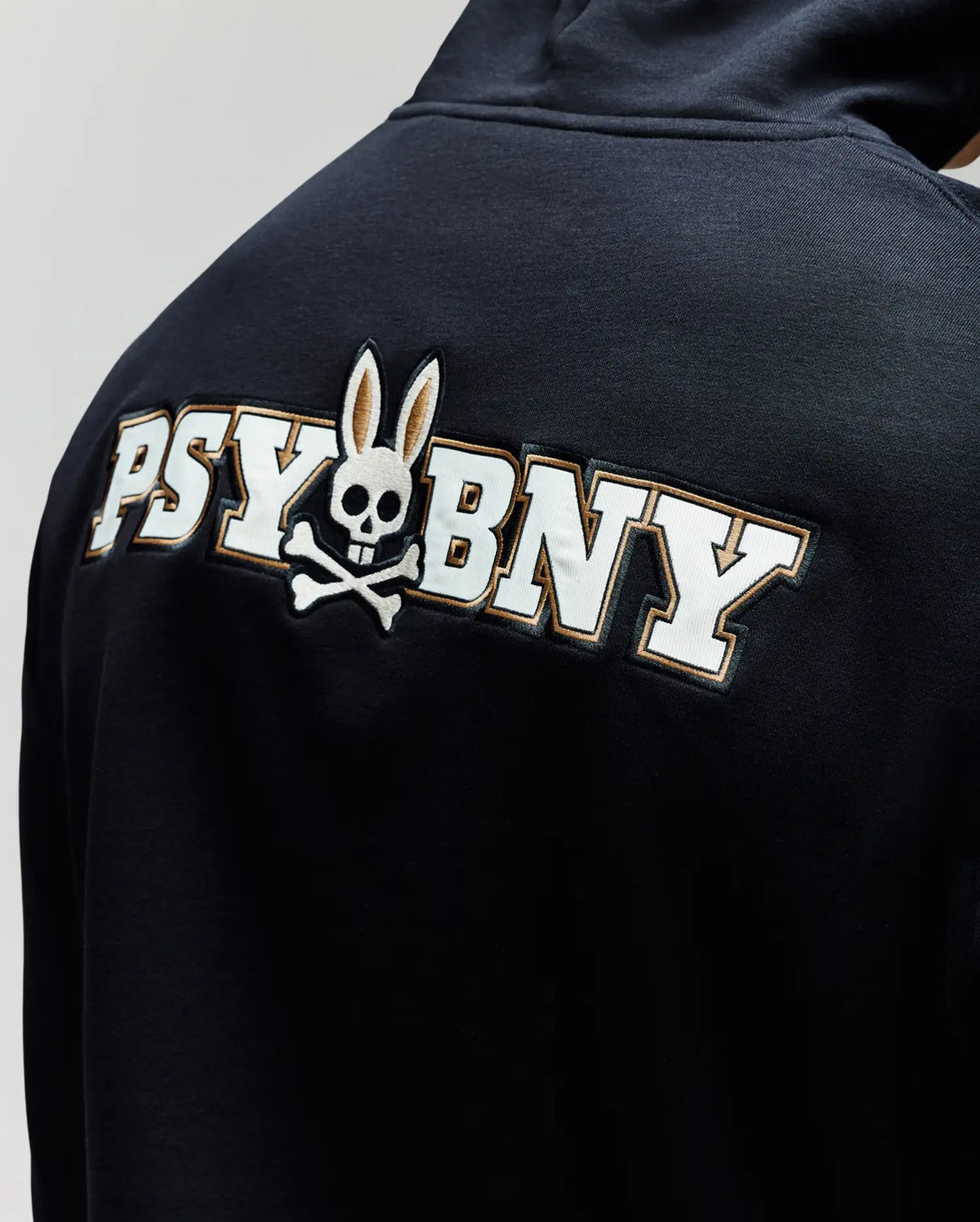 A close-up of the Psycho Bunny MENS WADE HOODIE (MT0500112) shows bold white "PSYBUNY" lettering on the back, featuring a bunny skull and crossbones in place of the "U"—all on a premium cotton blend.