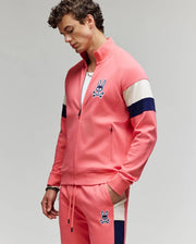 A man wears Psycho Bunny's MENS DAWSON TRACK JACKET - MT0500113 and matching joggers, both in pink with navy and white stripes, crafted from a soft cotton-modal blend with a rabbit skull logo. He stands looking down, one hand in his pocket.