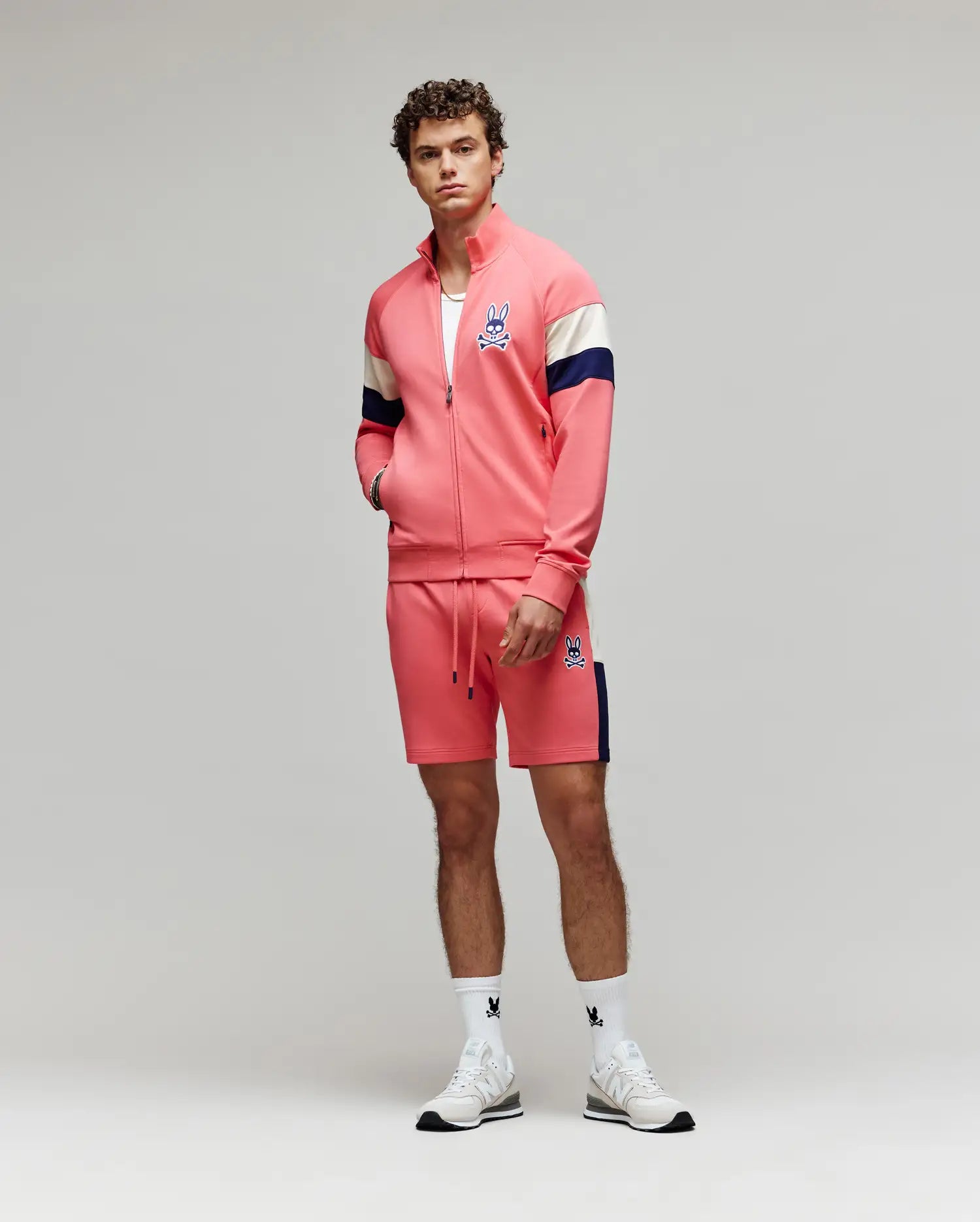 A young man stands facing forward in a Psycho Bunny MENS DAWSON TRACK JACKET (MT0500113) and matching shorts in coral pink with navy and white accents, paired with white socks and sneakers, set against a plain light gray background.