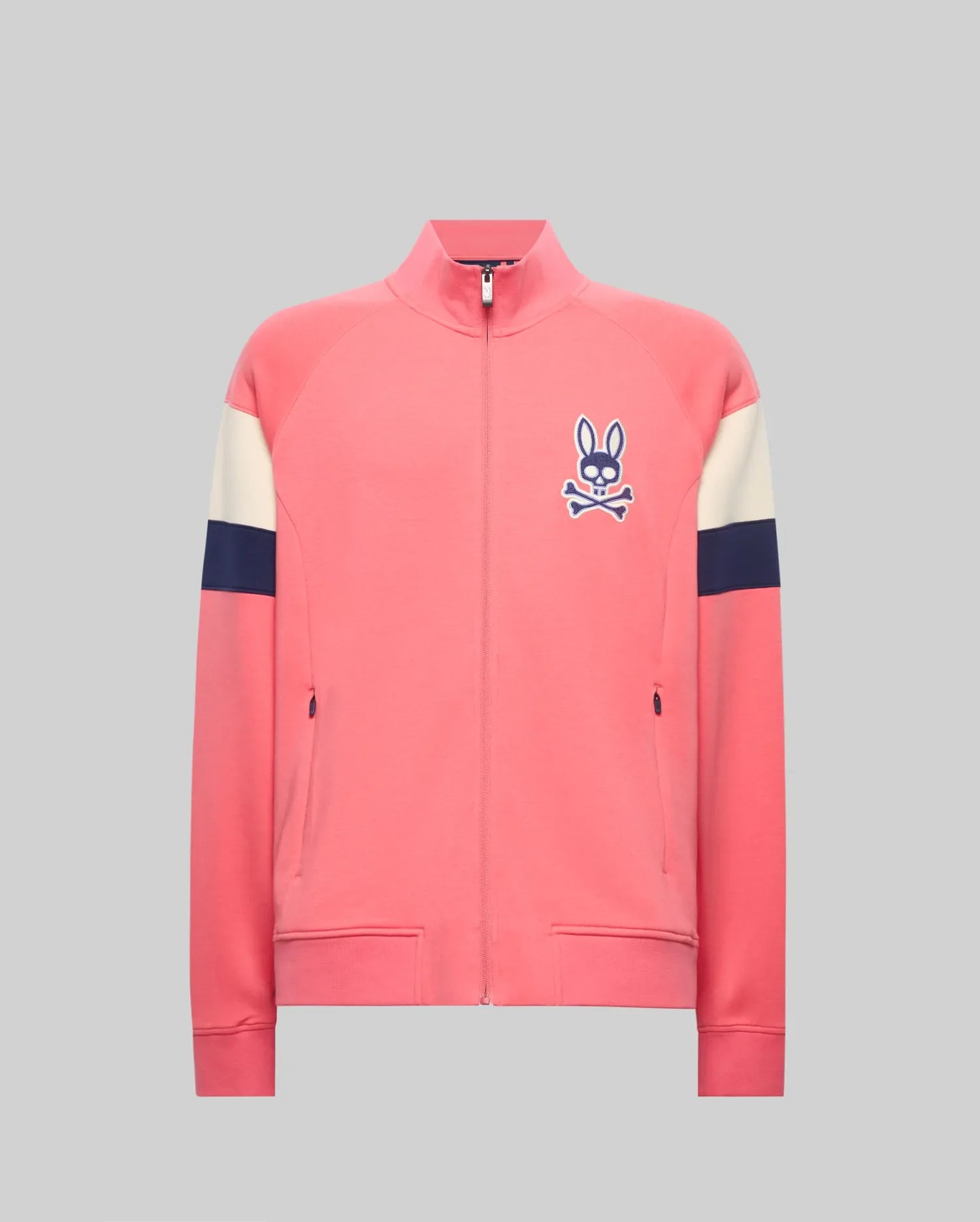 The Psycho Bunny MENS DAWSON TRACK JACKET (MT0500113) is a pink zip-up with cream and navy sleeve stripes, a blue bunny and crossbones logo on the left chest, made from a soft cotton-modal blend, shown on a plain gray background.