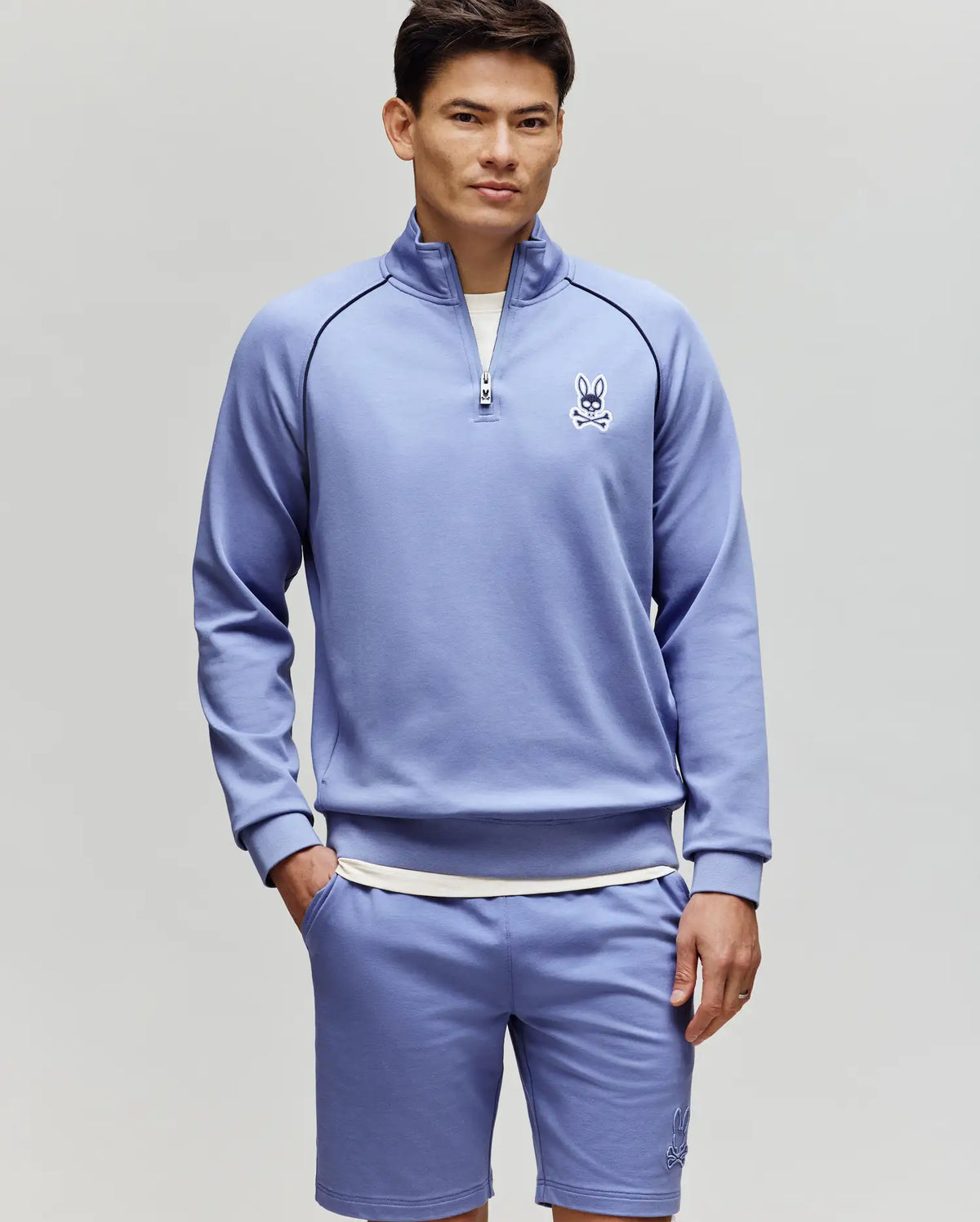 A man wears the Psycho Bunny MENS SHAWN QUARTER ZIP SWEATSHIRT - MT0500115 in light purple with matching shorts, both featuring a small bunny logo, standing against a plain background and looking at the camera with one hand in his pocket.