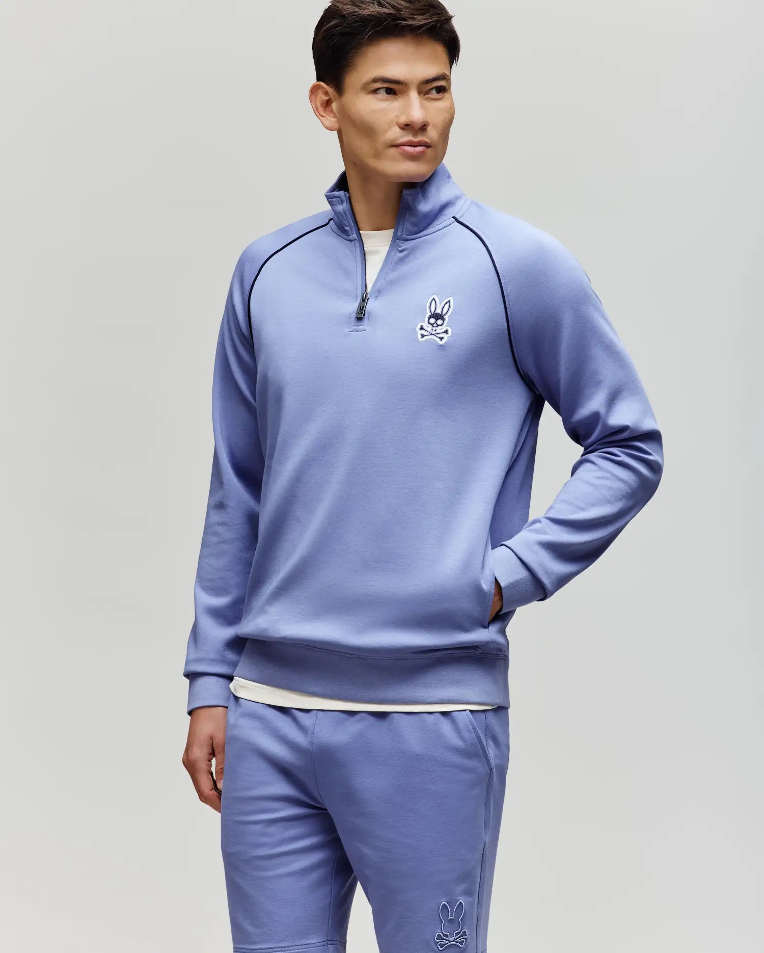 A man stands against a light gray background wearing the Psycho Bunny MENS SHAWN QUARTER ZIP SWEATSHIRT (MT0500115) in light purple with matching white bunny logo shorts, looking to the side with his hands in his pockets.