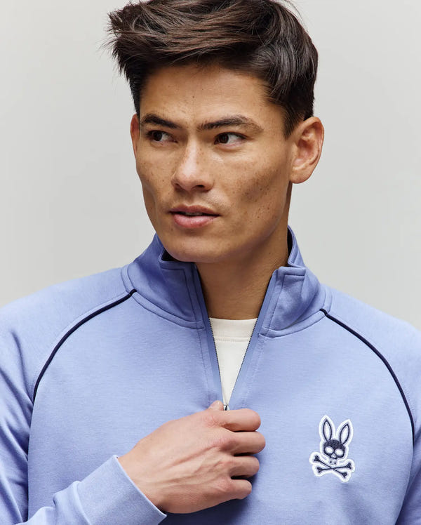 A man in the Psycho Bunny MENS SHAWN QUARTER ZIP SWEATSHIRT (MT0500115), made from premium cotton-modal knit, wears a light blue top with a white bunny and crossbones chest logo, looking to the side while holding the 1/4 zip.
