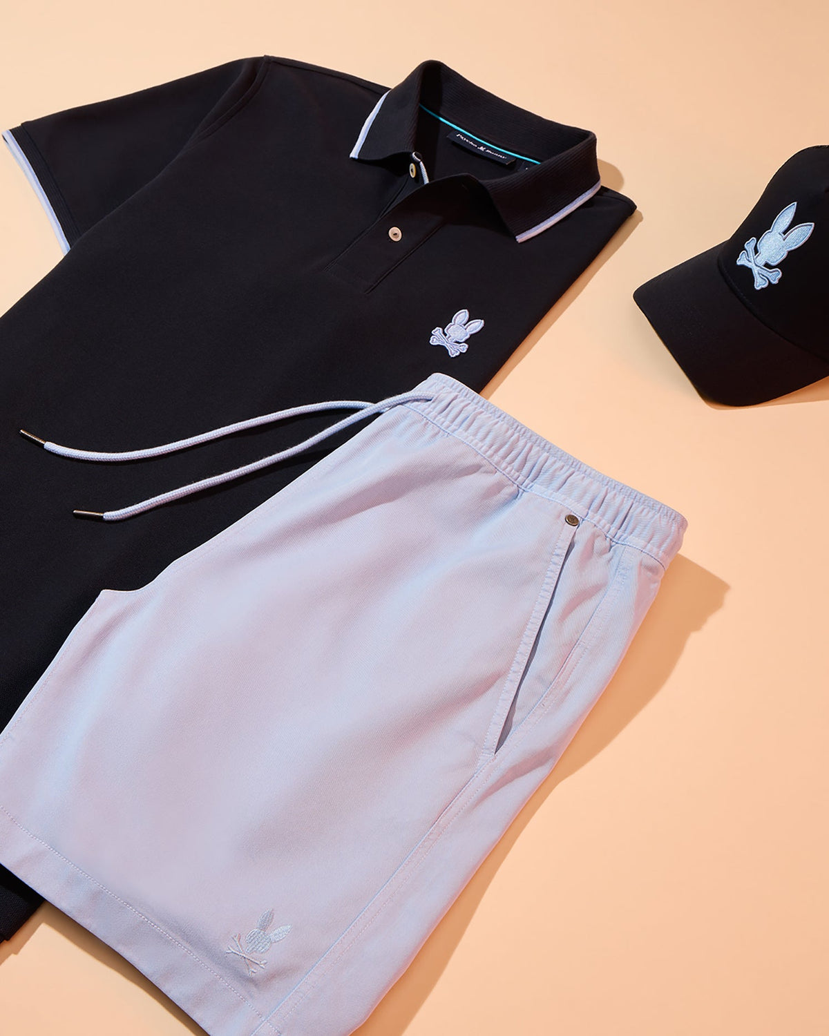 A stylish ensemble featuring a navy polo shirt, light pink Psycho Bunny MENS WILLIS STRETCH TENCEL SHORT - B6R239Y1WB, and a black baseball cap, all embroidered with a white floral motif, displayed on a peach background.