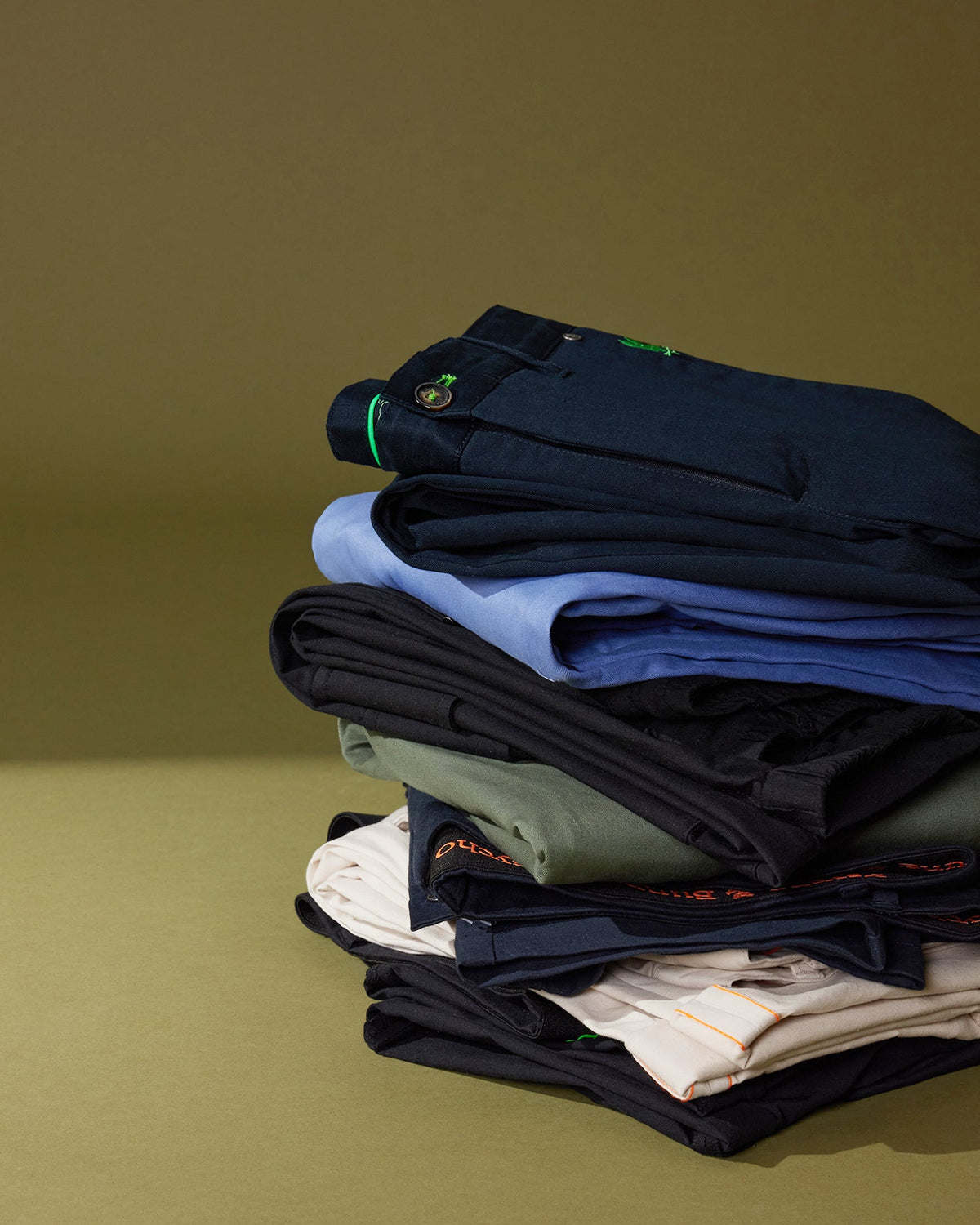 A neatly stacked pile of various pieces of clothing, including Psycho Bunny men's Gable regular fit sport pants and shirts, on a green background. The clothes feature subtle, stylish stitching and detailing.