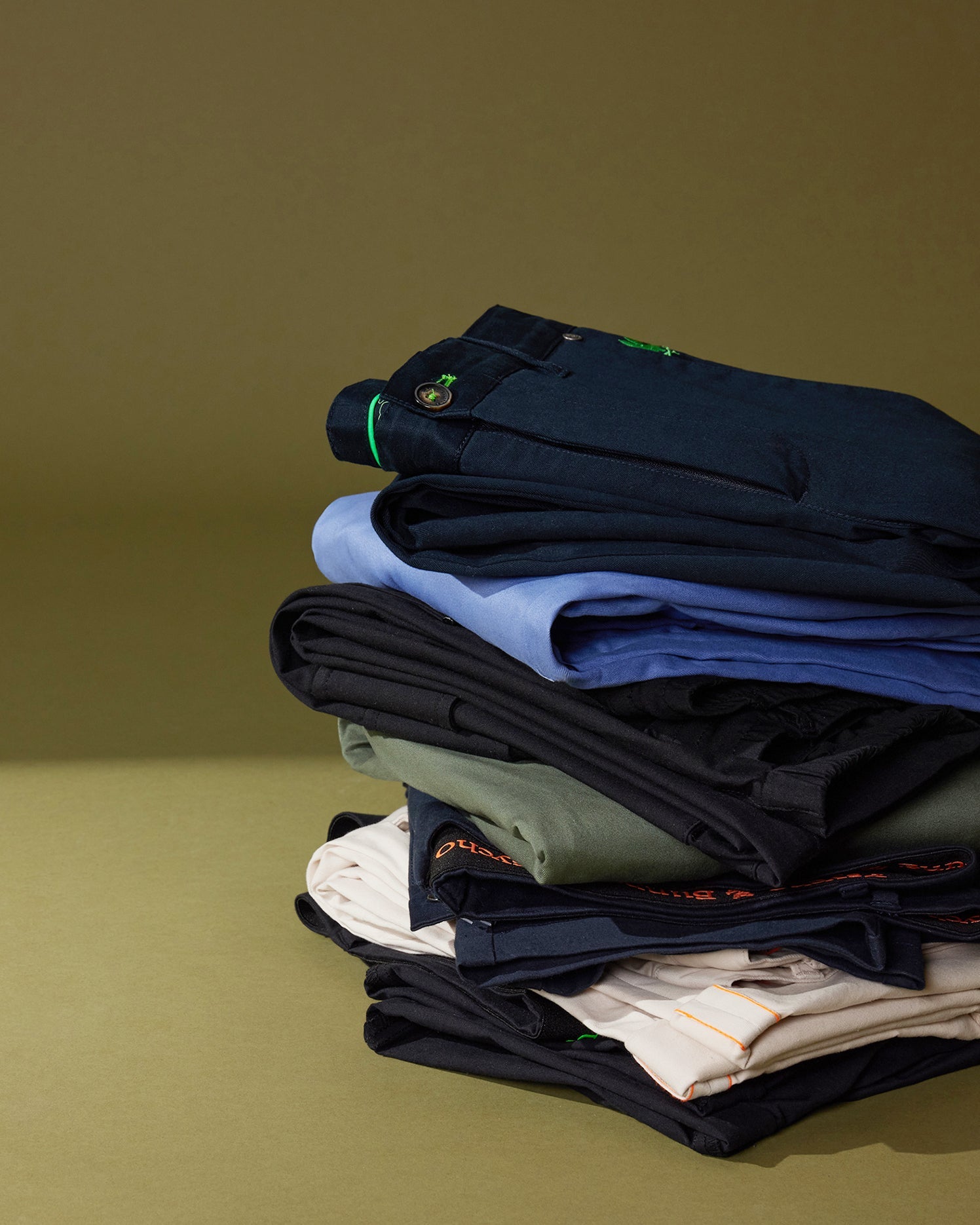 A neatly stacked pile of various pieces of clothing, including Psycho Bunny men's Gable regular fit sport pants and shirts, on a green background. The clothes feature subtle, stylish stitching and detailing.