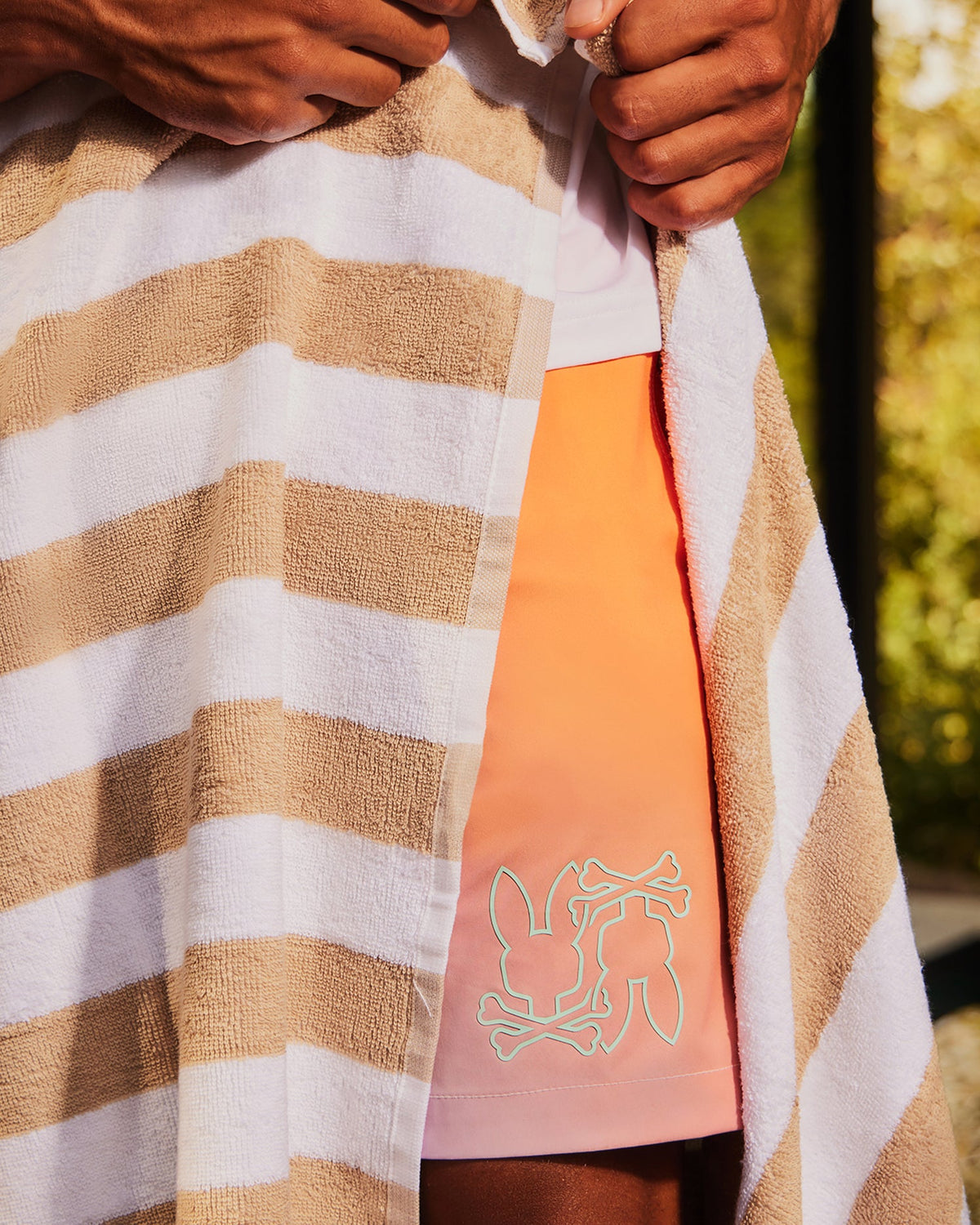 A person wrapped in a brown and white striped towel reveals part of their outfit, which includes a pair of Psycho Bunny MENS WINTON PRINTED SWIM TRUNK - B6W915A2PO. The background shows a blurred outdoor setting with greenery.