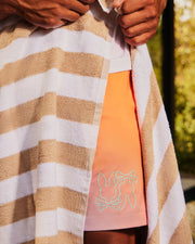 A person wrapped in a brown and white striped towel reveals part of their outfit, which includes a pair of Psycho Bunny MENS WINTON PRINTED SWIM TRUNK - B6W915A2PO. The background shows a blurred outdoor setting with greenery.