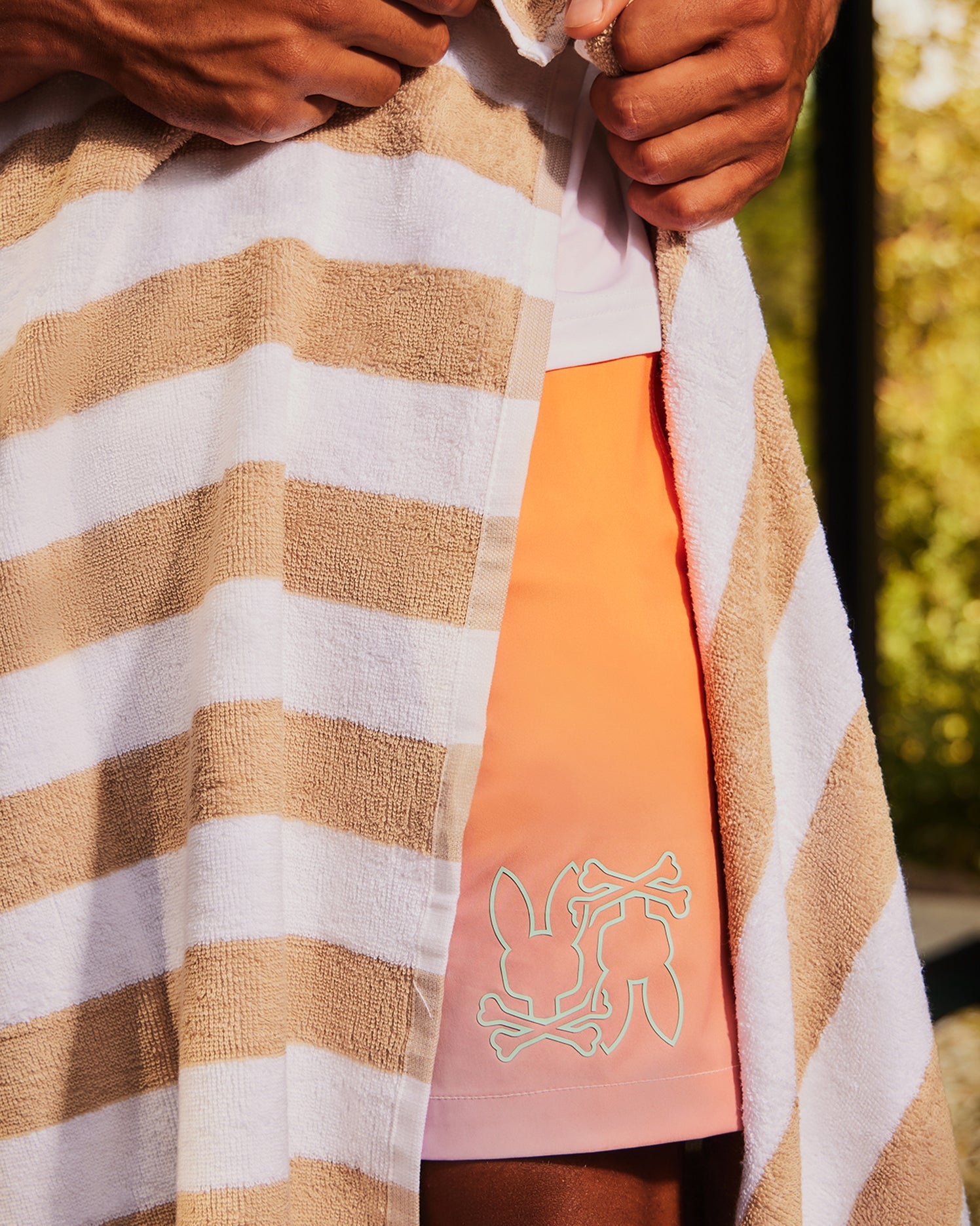 A person wrapped in a brown and white striped towel reveals part of their outfit, which includes a pair of Psycho Bunny MENS WINTON PRINTED SWIM TRUNK - B6W915A2PO. The background shows a blurred outdoor setting with greenery.